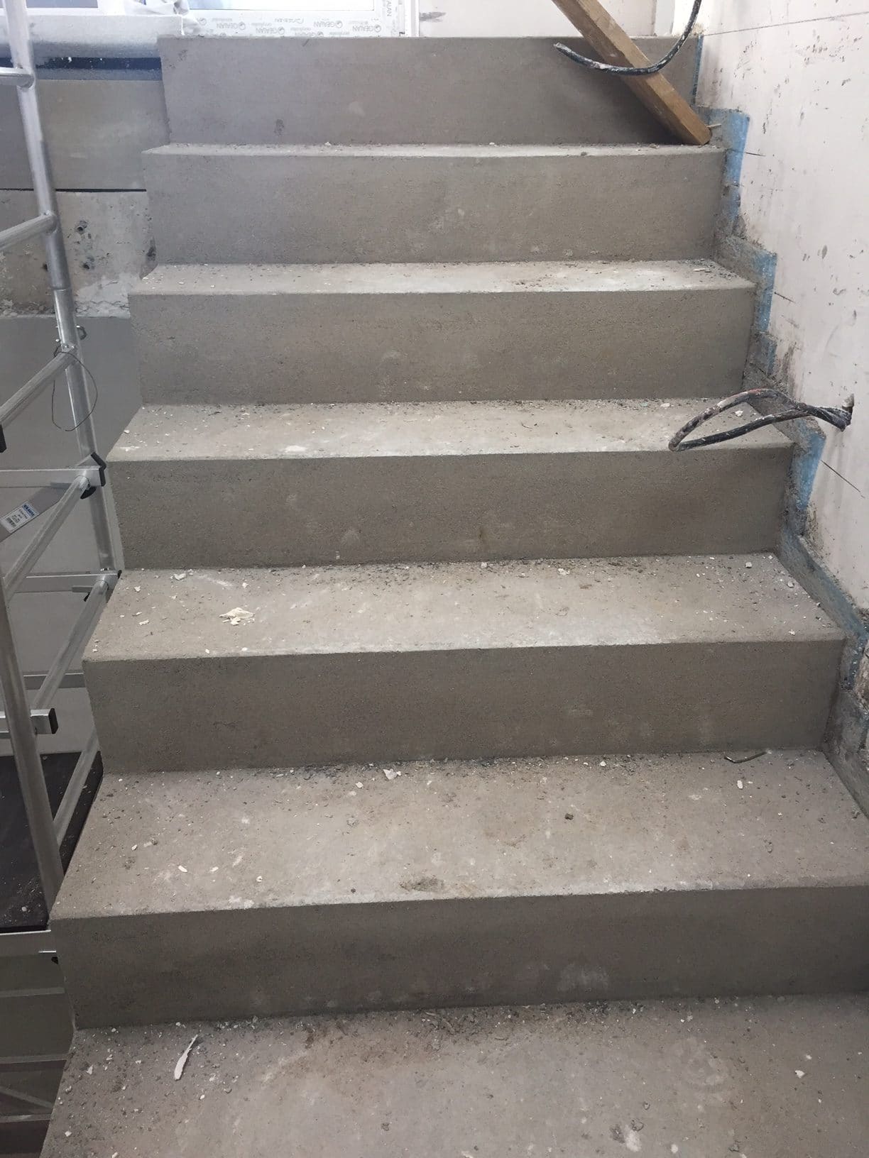 Eine unvollendete Treppe aus grauem Beton, die mit Staub und Bauschutt bedeckt ist. Im Hintergrund sind verputzte Wände und ein Gerüst zu sehen.