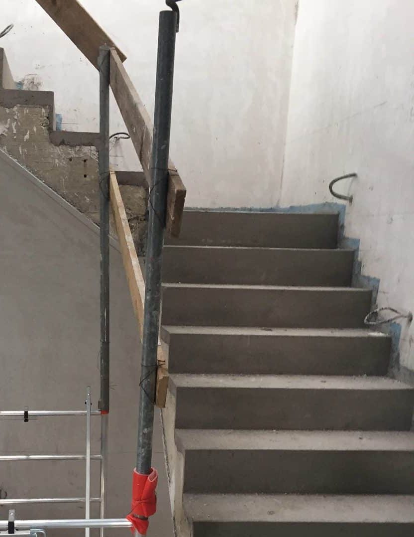 Eine unbehandelte Treppe mit grauen Betonstufen und einem einfachen Holzgeländer. An der Seite ist ein Metallgerüst zu sehen, das für Bauarbeiten verwendet wird. Die Wände sind unvollständig verputzt und zeigen Baustellenmerkmale.