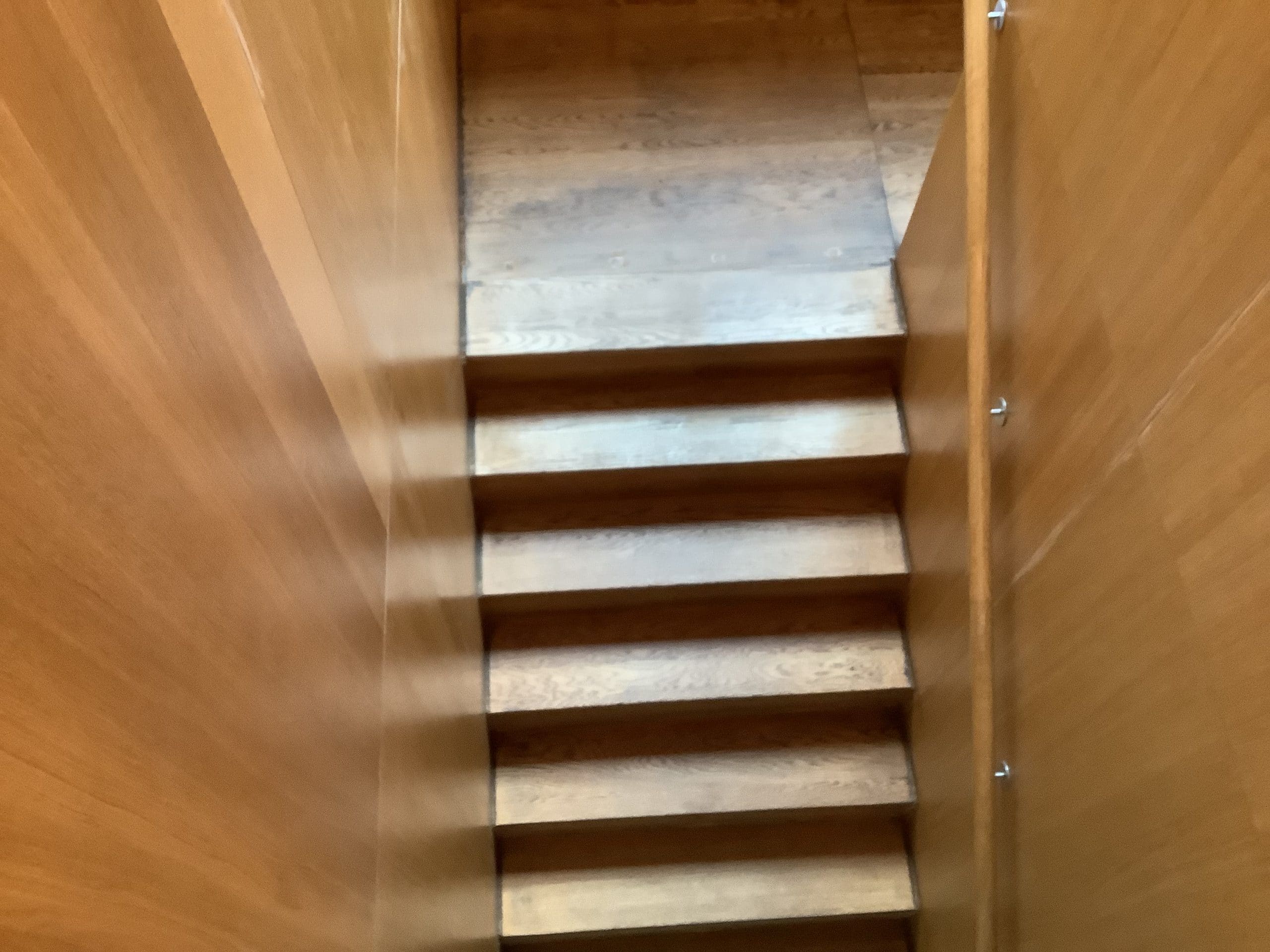 Eine Treppe aus Holz, die nach unten führt, umgeben von hellen Holzpaneelen. Das Licht fällt auf die Stufen, die gleichmäßig angeordnet sind.