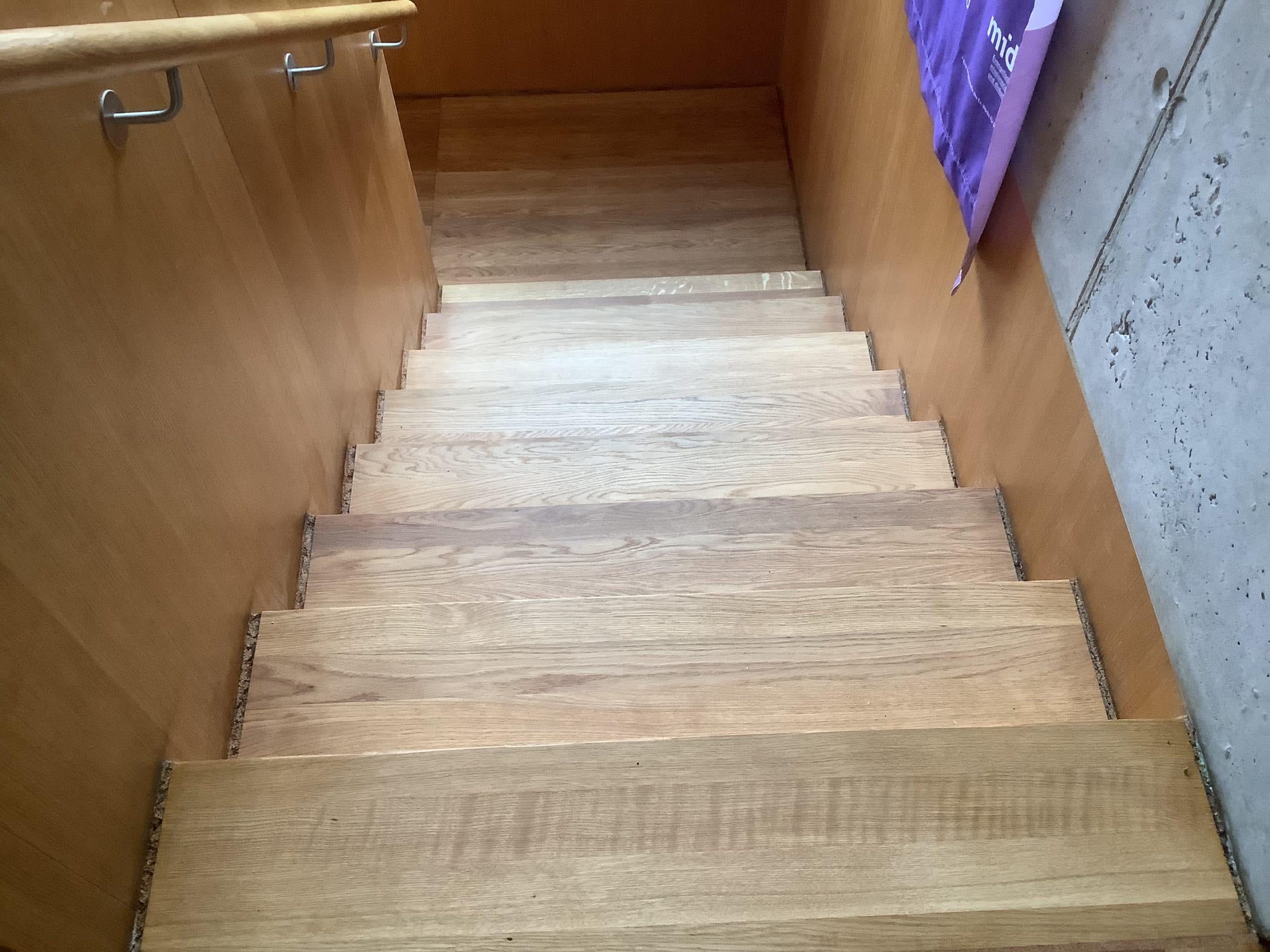 Eine Treppe mit hellen Holzstufen, die zu einem unteren Bereich führt. An der Wand sind helle Holzverkleidungen und ein grauer Betonabschnitt sichtbar. Ein Handlauf aus Metall verläuft entlang der Treppe.