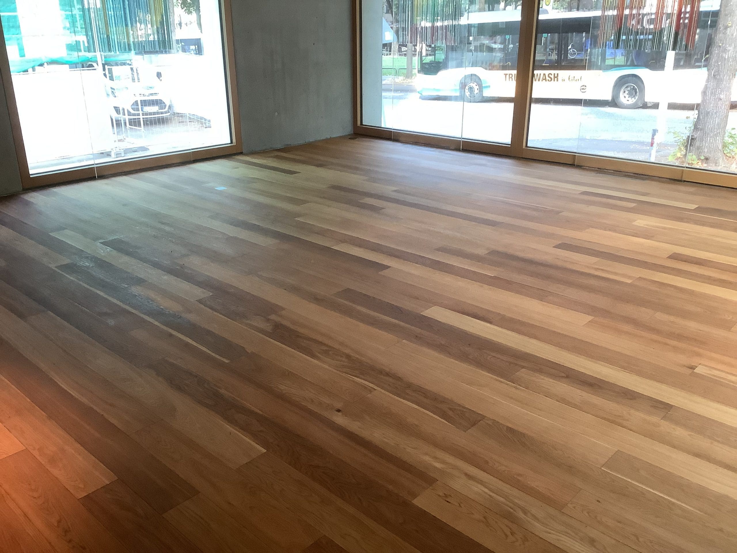 Ein heller Holzfußboden mit verschiedenen Holzarten und -tönen, der in einem Raum mit großen Fenstern und einem minimalistischen, modernen Ambiente verlegt ist. Im Hintergrund sind eine Straße und ein Bus sichtbar.
