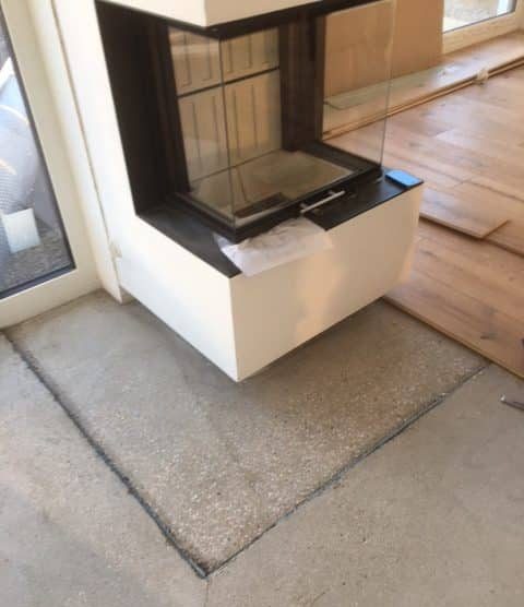 Moderne Kamininstallation in einem Raum mit einem gläsernen Sichtfenster und einer weißen, minimalistischen Umrandung. Der Boden ist teilweise mit Rohbeton und Holzfußboden verlegt.
