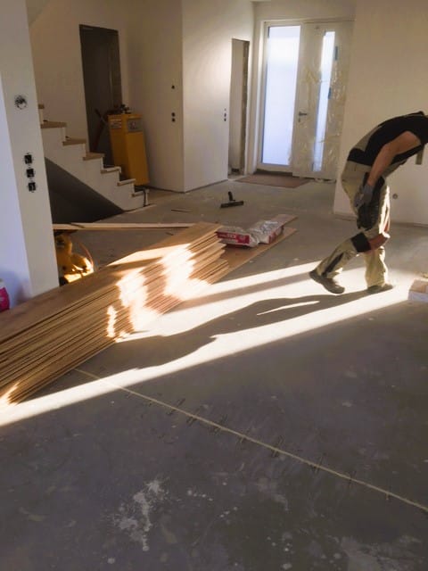 Ein Handwerker arbeitet in einem unvollendeten Raum, in dem Holzplatten auf dem Boden liegen. Im Hintergrund sind Treppen und eine große Fensterfront zu sehen, durch die Licht strömt.