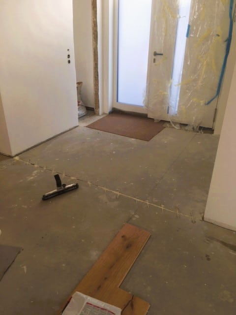 Ein unvollendeter Flur mit einem Sicht auf eine Eingangstür. Der Boden besteht aus rohem Beton, und einige Holzplanken liegen verstreut. An der Wand sind Reste von Bauarbeiten sichtbar, während die Tür mit einer Plastikfolie abgedeckt ist.