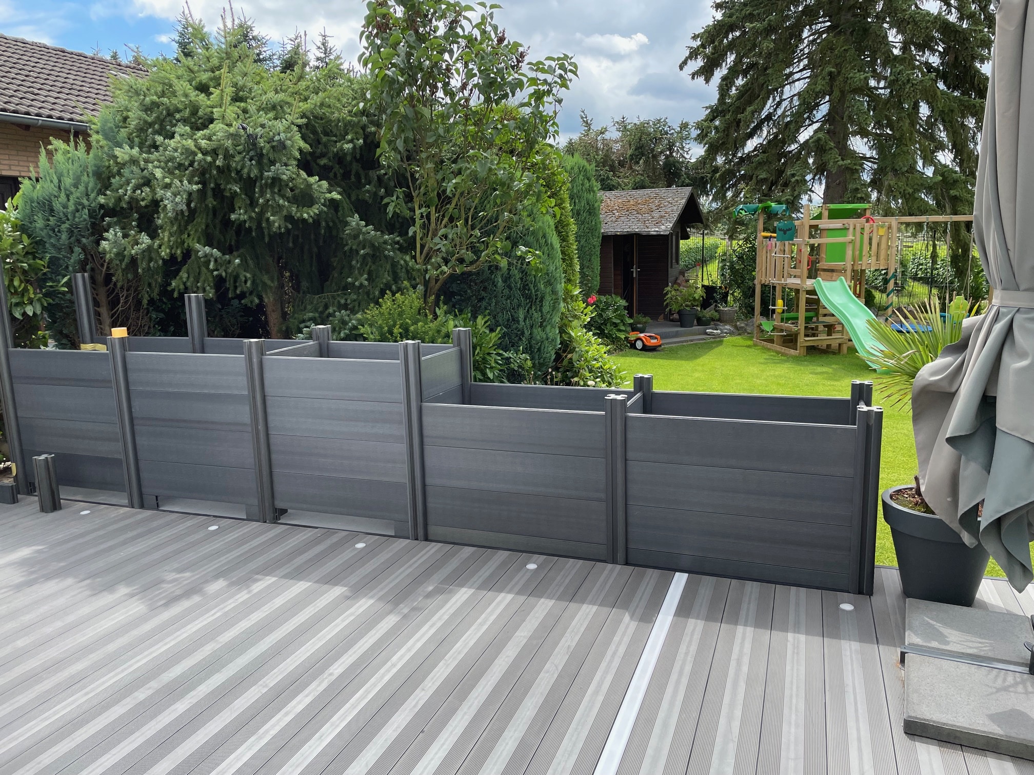 Ein Gartenbereich mit einem grauen Sichtschutzzaun, einer Holzterrasse und grünen Pflanzen im Hintergrund. Im Hintergrund sind ein Spielplatz mit Rutsche und ein kleines Gartenhaus zu sehen.