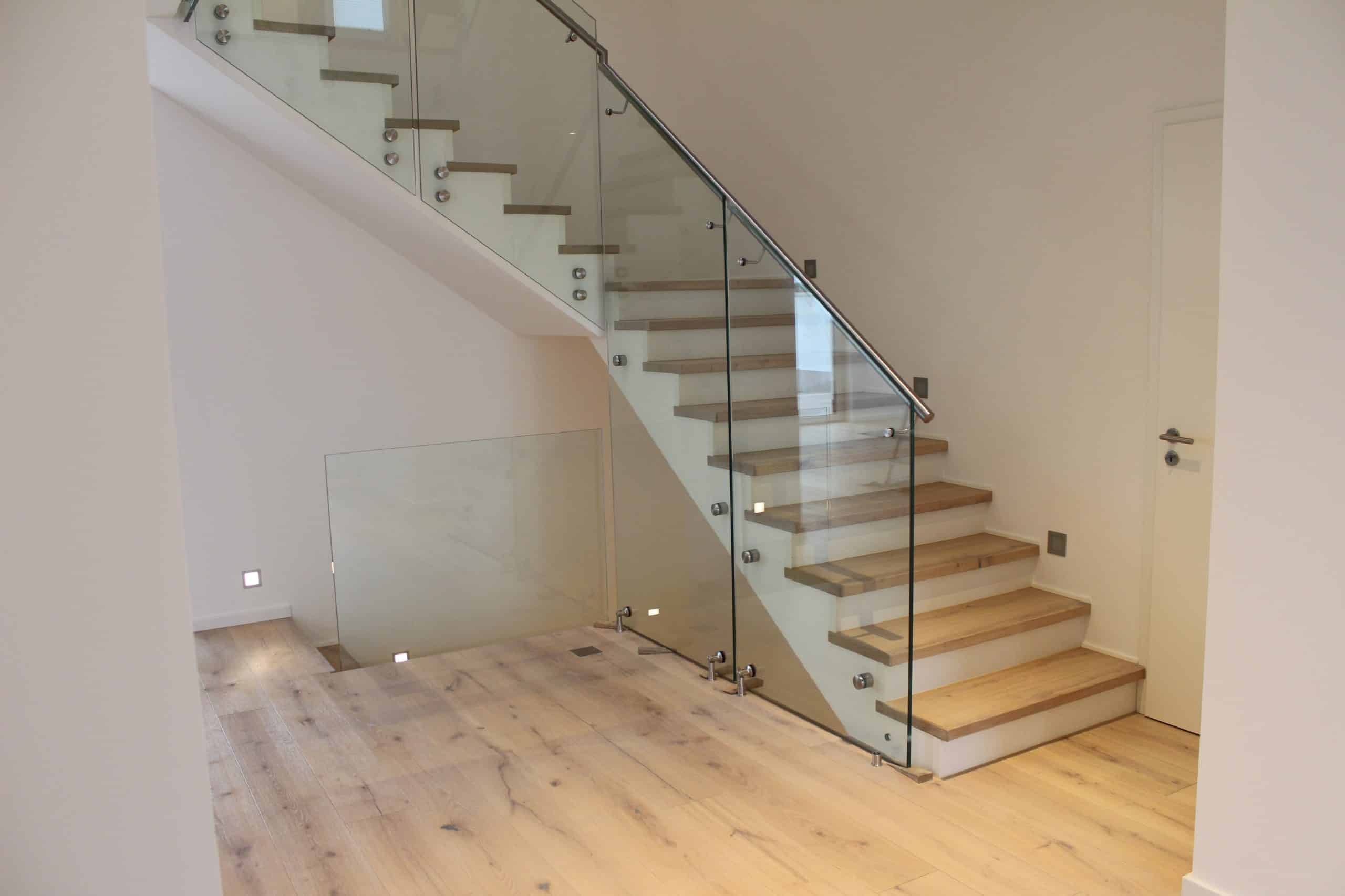 Ein moderner Treppenaufgang mit Holzstufen und einer transparenten Glasgeländer. Der Raum hat helle Wände und einen warmen Holzboden, der eine einladende Atmosphäre schafft.