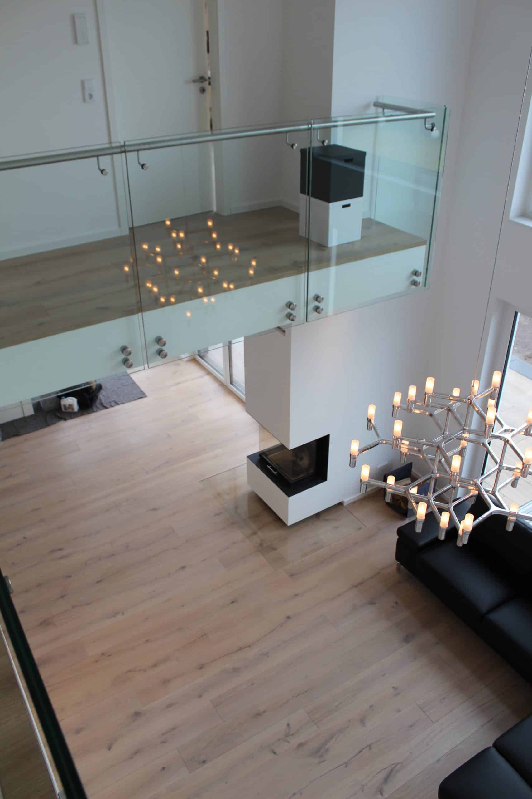 Ein moderner Wohnraum mit Holzfußboden, einer eleganten Glaskonstruktion im Obergeschoss und einem stilvollen Kronleuchter. Im Hintergrund ist ein minimalistischer Kamin zu sehen, während ein schwarzes Sofa den Raum ergänzt.
