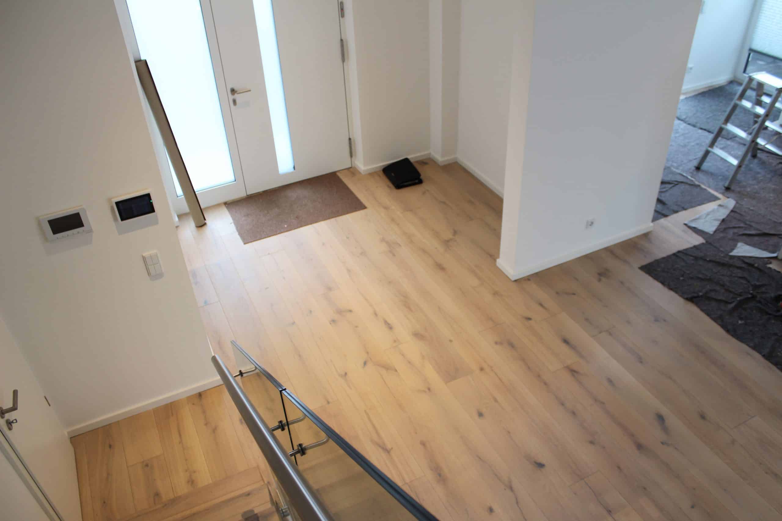 Der Eingangsbereich eines modernen Hauses mit hellen Holzfußböden, einer weißen Wand und einer großen Glastür, die Tageslicht hereinlässt. An der Wand sind zwei Steuergeräte angebracht, und ein Teppich liegt vor der Tür. Im Hintergrund sind einige Baustellenutensilien sichtbar.