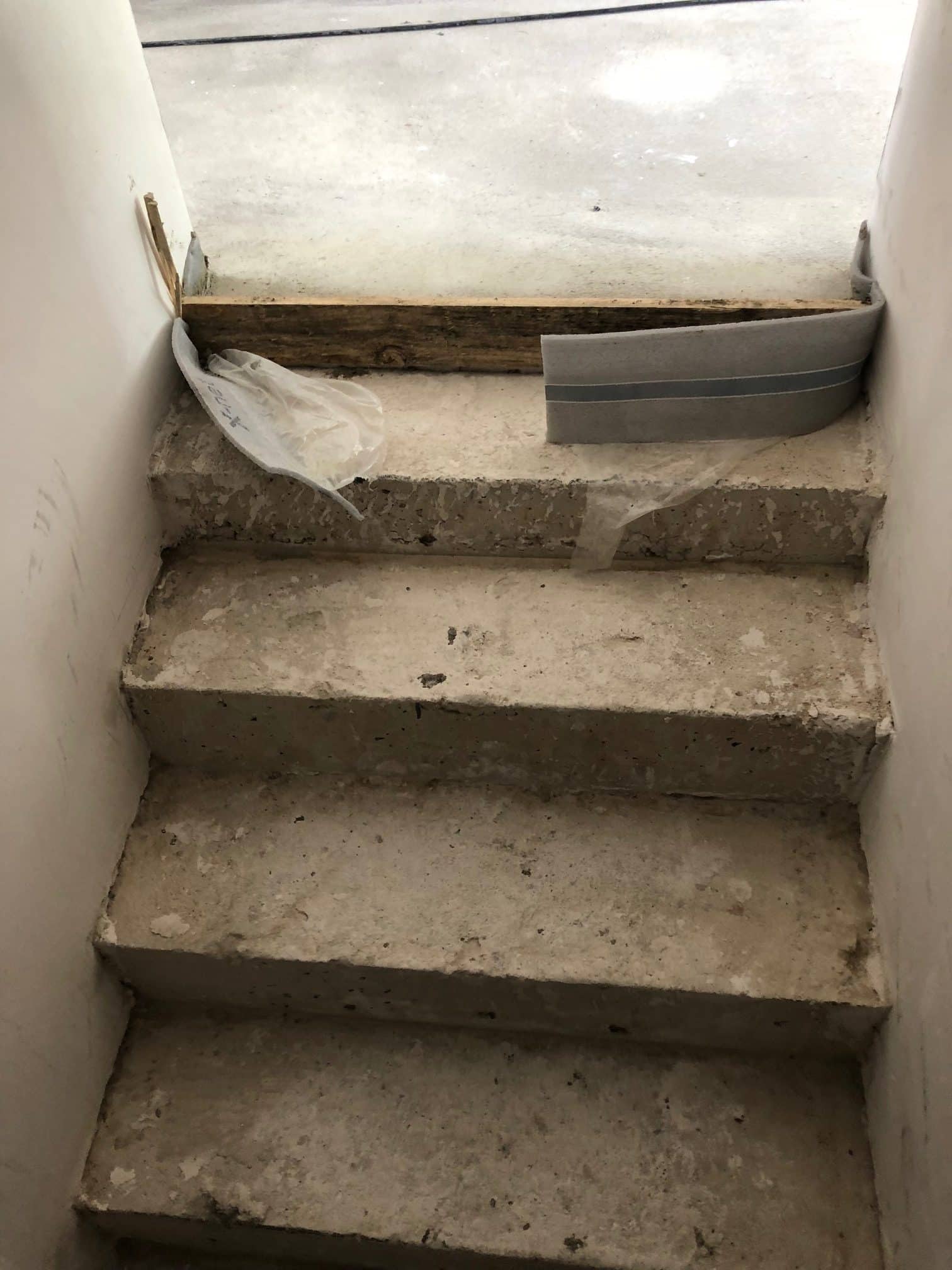 Eine Treppe aus rohem Beton, die leicht abgenutzt ist, führt nach unten. An der Wand sind einige Reste von Bauarbeiten zu sehen, darunter ein Stück Holz und ein grauer Streifen. Es liegt ein Stück Plastikfolie auf den Stufen. Der Boden oben ist uneben und teilweise sichtbar.
