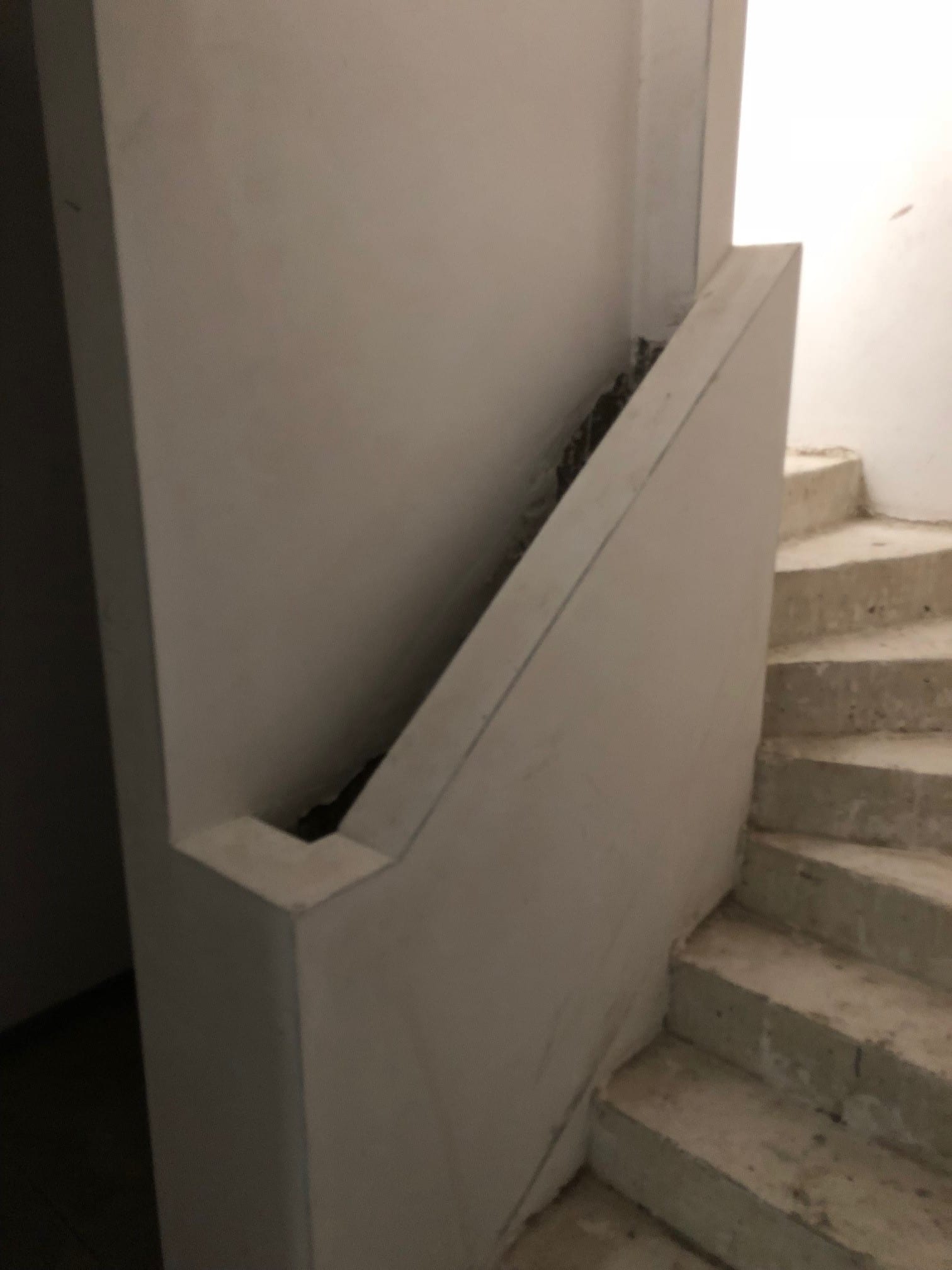 Eine schlichte Treppe in einem Innenraum, die aus grauem Beton besteht. An der Seite befindet sich eine weiße Wand mit einer schrägen Kante, die den Treppenaufgang begrenzt. Der Raum wirkt minimalistisch und nüchtern, mit gedämpften Lichtverhältnissen.