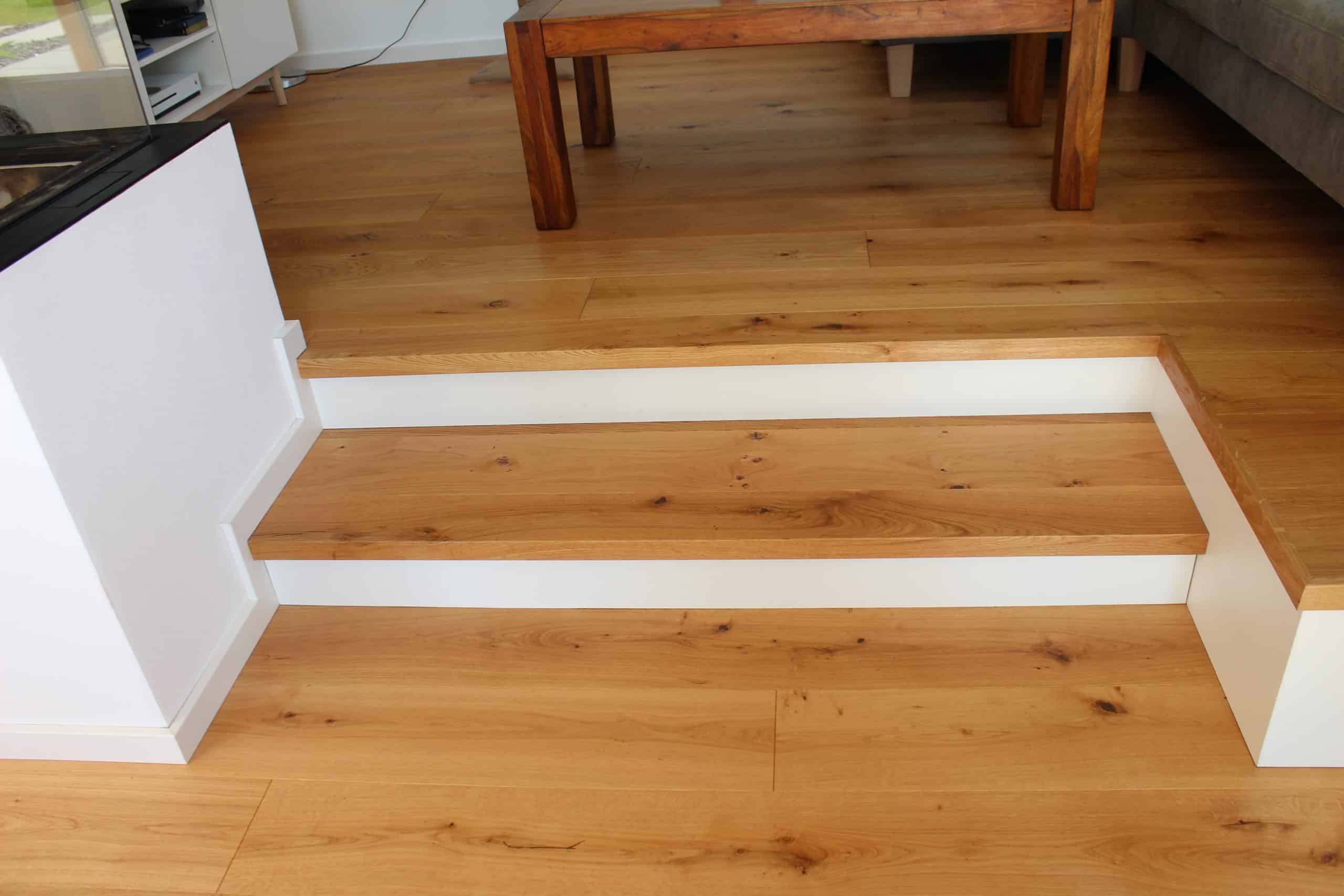 Eine Treppe mit Holzstufen und weißen Wangen, die in einen Raum mit einem hellen Holzfußboden führt. Im Hintergrund ist ein Tisch aus massivem Holz zu sehen.