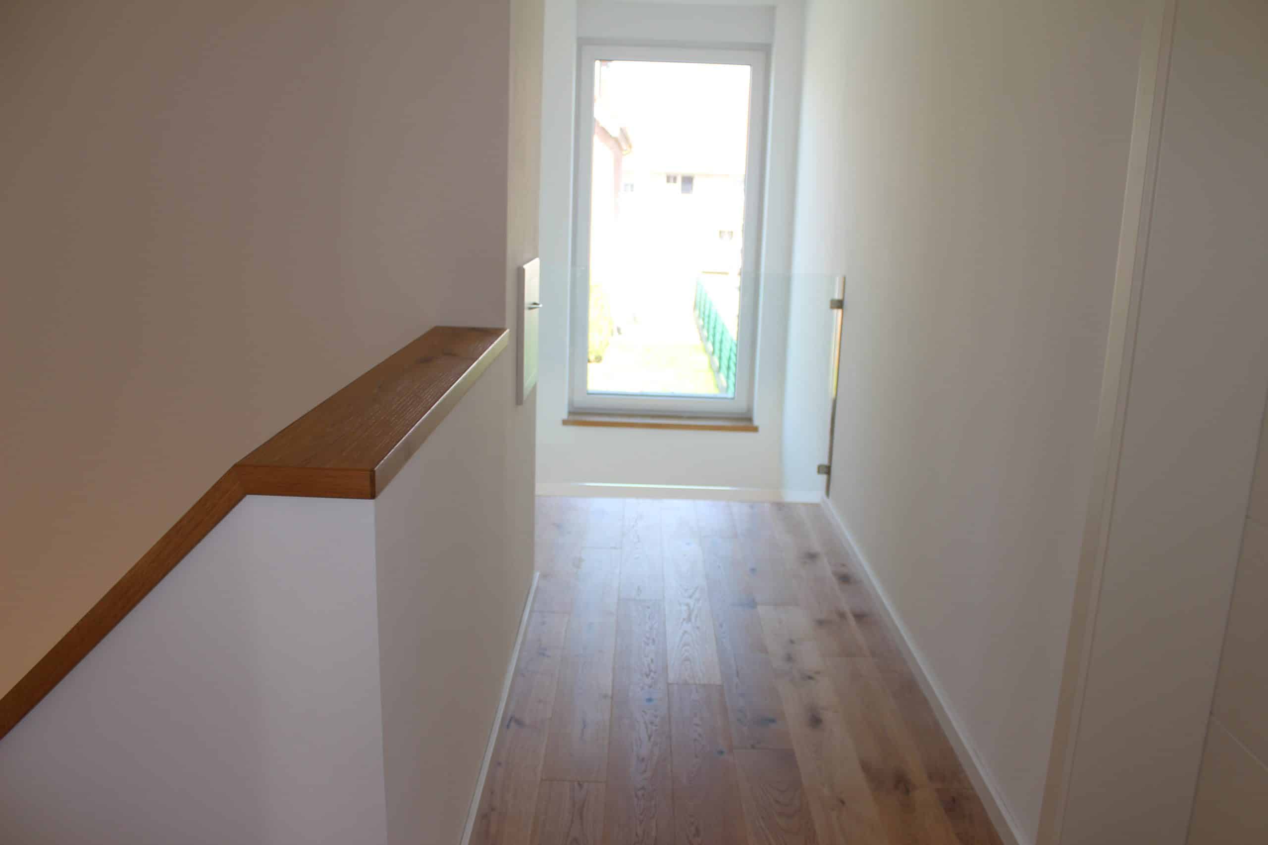 Ein heller Flur mit Holzfußboden und einem Geländer aus Holz, das an einer weißen Wand verläuft. Am Ende des Flurs befindet sich ein großes Fenster, das Tageslicht hereinlässt und auf einen Außenbereich blickt.