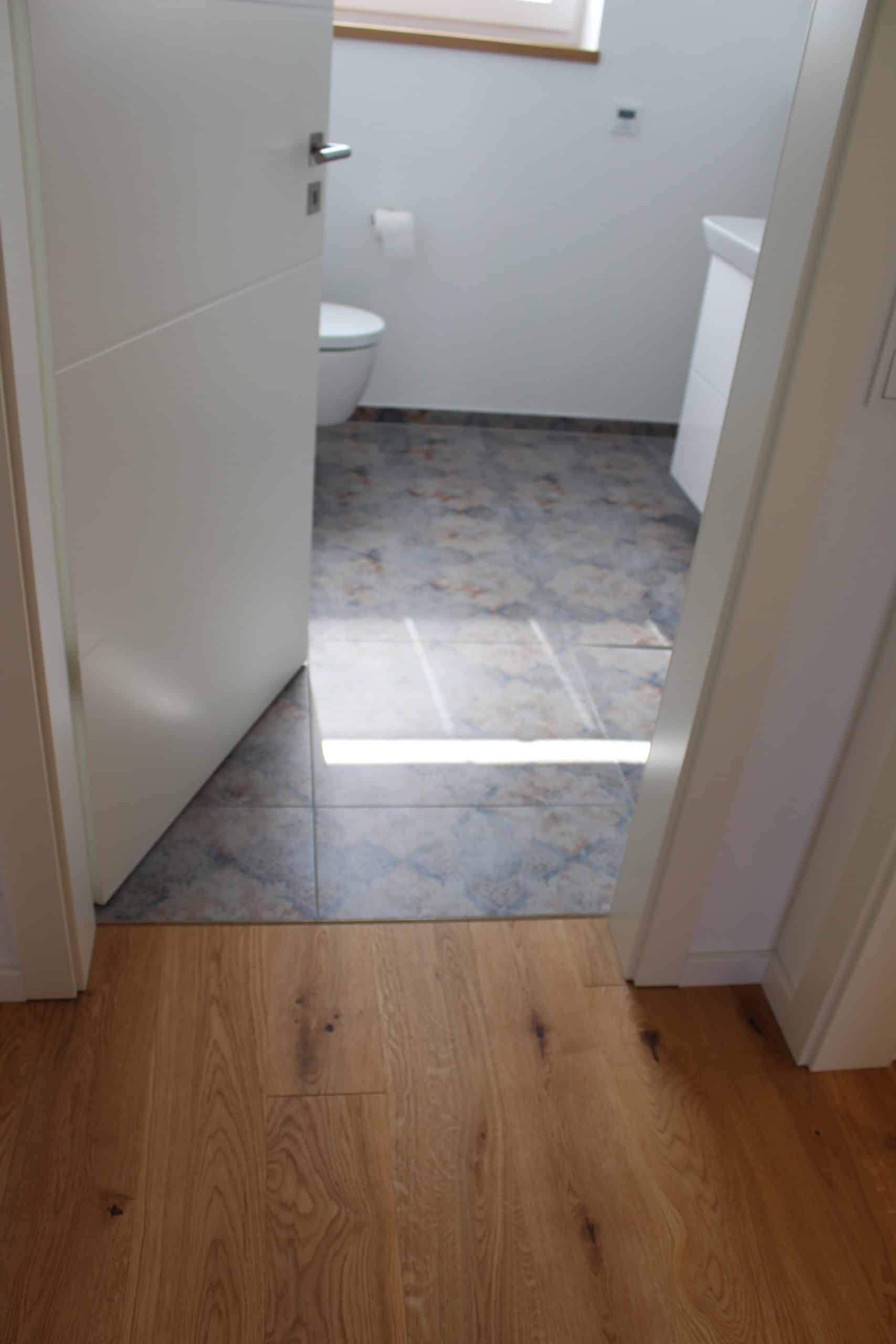 Blick durch eine offene Tür auf einen modernen Badezimmerboden, der mit grauen Fliesen und hellem Holzparkett gestaltet ist. Im Hintergrund ist eine Toilette und ein Waschbecken sichtbar, während Lichtstrahlen den Raum erhellen.
