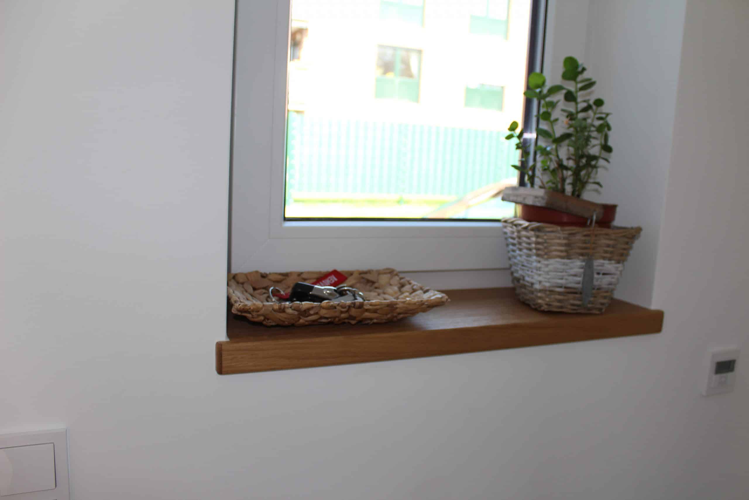 Fensterbank mit einem geflochtenen Korb, in dem sich Schlüssel befinden, und einem Topf mit einer kleinen Pflanze. Im Hintergrund ist ein Fenster sichtbar, das auf einen Außenbereich blickt.