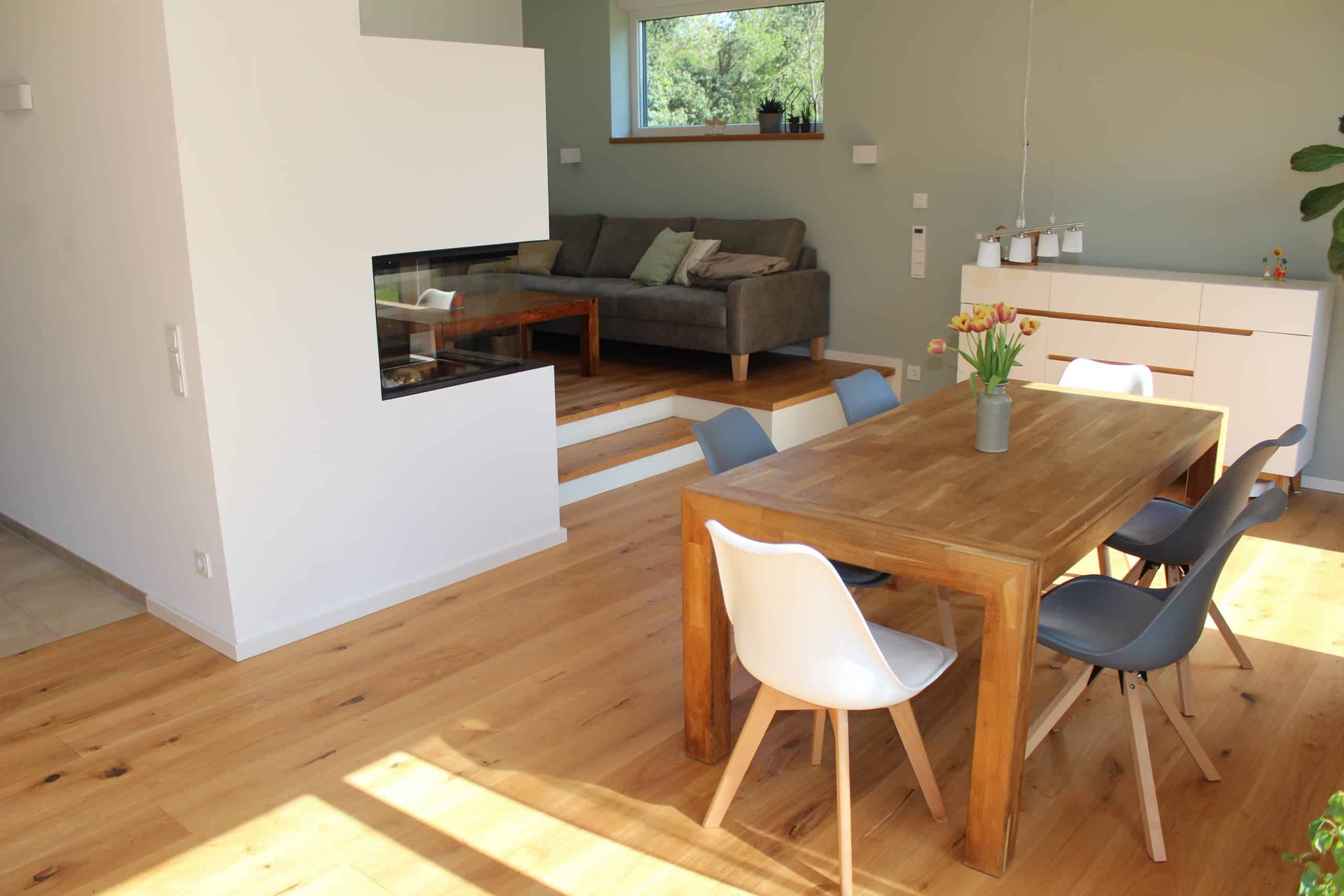 Ein modernes Wohnzimmer mit einem Holzfußboden, einem Esstisch aus Holz und Stühlen in verschiedenen Farben. Im Hintergrund befindet sich eine gemütliche Couch und ein Kamin in einer weißen Wand. Große Fenster lassen viel Tageslicht herein.