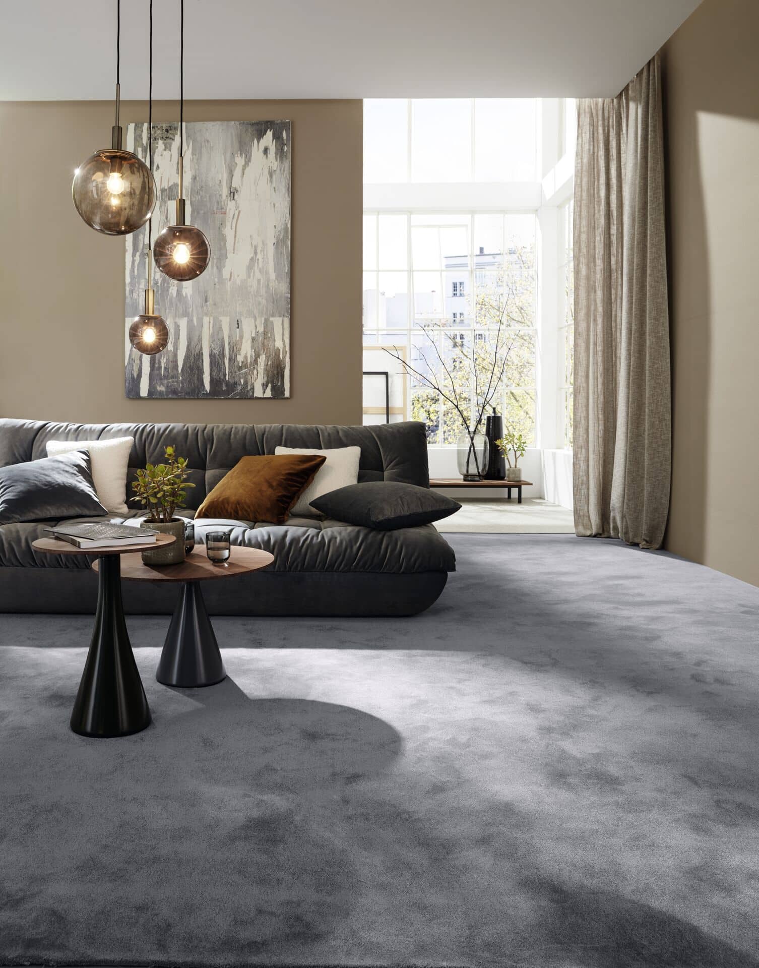 Ein stilvolles Wohnzimmer mit einem grauen Teppich, einer bequemen, dunklen Couch mit bunten Kissen, einem kleinen Tisch mit einer Pflanze und modernen Pendelleuchten. Große Fenster lassen viel Tageslicht herein und schaffen eine helle Atmosphäre.