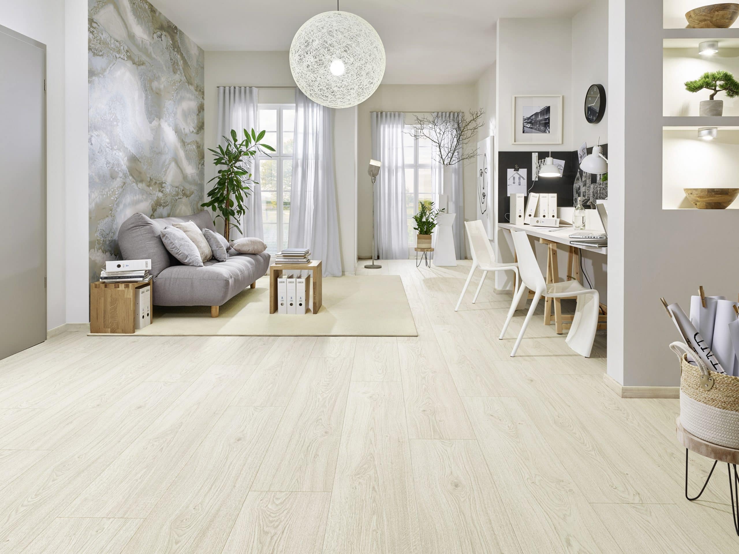 Ein modernes, helles Wohnzimmer mit einem grauen Sofa, Holzregalen und einem Schreibtischbereich. Große Fenster lassen viel Licht herein, und eine dekorative Lampe hängt von der Decke. Die Wände sind in sanften Farben gehalten, mit einer auffälligen Tapete und Pflanzen zur Dekoration.