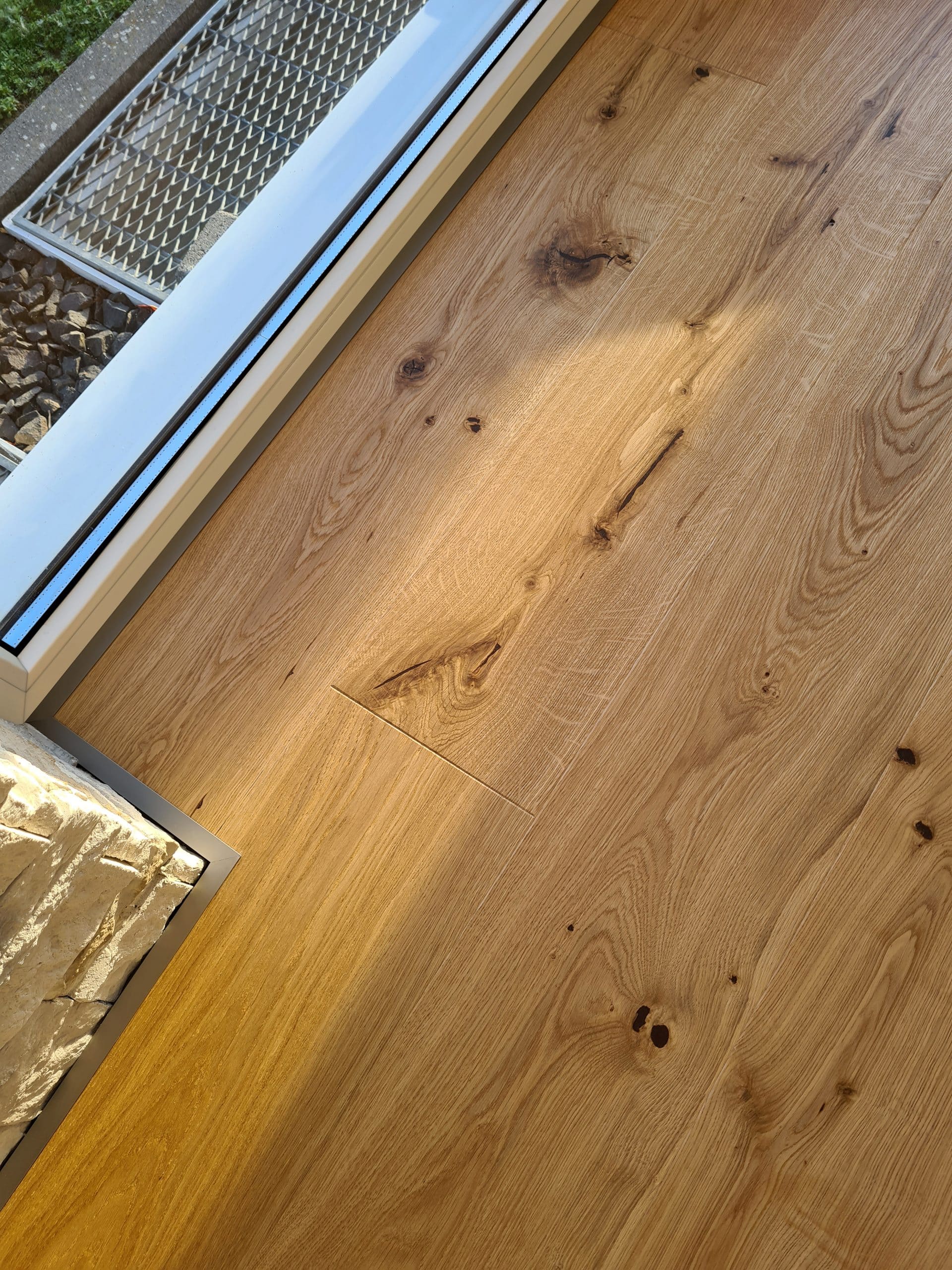 Ein heller Holzboden aus Eiche mit sichtbaren Maserungen und kleinen Ästen, der in einem Raum verlegt ist. Im Hintergrund ist ein Fensterrahmen und ein Teil eines Gartenbereichs zu sehen.