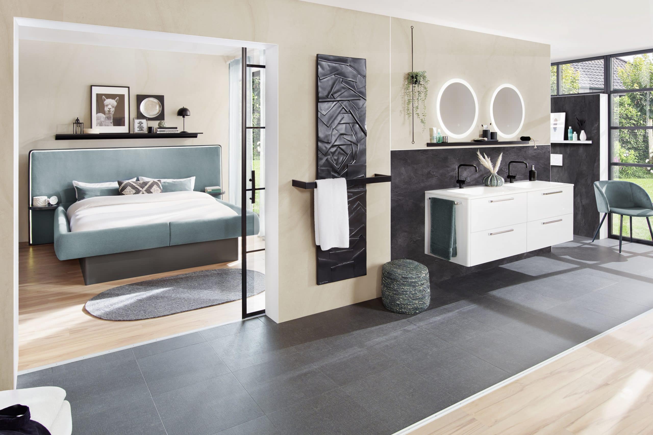 Ein modernes Schlafzimmer mit einem gepolsterten Bett in sanften Farbtönen, einem runden Teppich und einer offenen Verbindung zu einem stilvollen Badezimmer. Das Badezimmer verfügt über eine elegante Doppelsenke, schwarze Armaturen und runde Spiegel, ergänzt durch eine Wand mit dekorativen Elementen und einem Handtuchhalter. Große Fenster sorgen für viel Tageslicht.