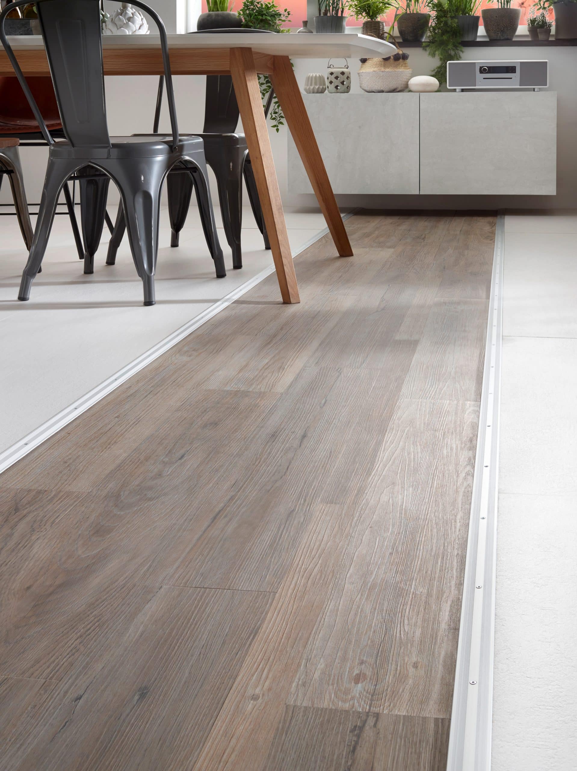 Der Boden eines modernen Essbereichs mit einem Holzlook, der sich unter einem Tisch mit Holzbeinen und Metallstühlen erstreckt. Im Hintergrund sind Pflanzen und ein Sideboard sichtbar, das eine dekorative Atmosphäre schafft.