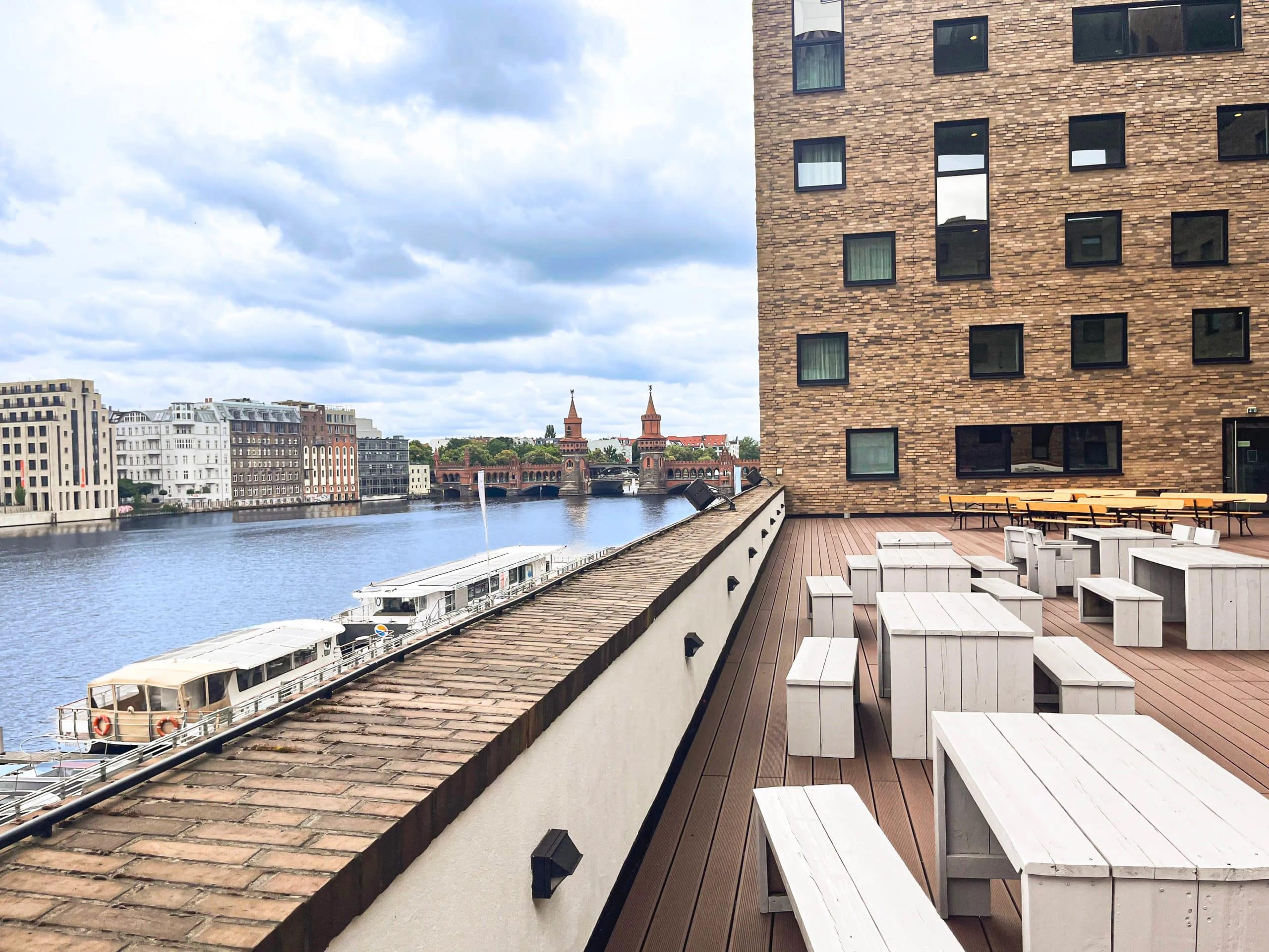 Blick von einer Terrasse auf einen Fluss, umgeben von modernen und historischen Gebäuden. Im Vordergrund stehen weiße Holzmöbel, während im Hintergrund eine Brücke mit Türmen sichtbar ist. Der Himmel ist bewölkt und das Wasser spiegelt die Umgebung wider.