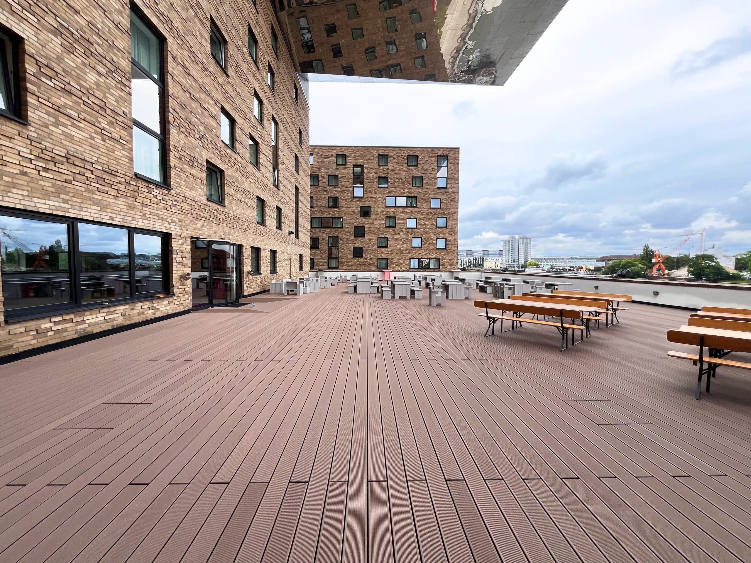 Eine große, moderne Dachterrasse mit Holzbelag, umgeben von einem Gebäude mit Backsteinfassade. Auf der Terrasse stehen mehrere Tische und Bänke sowie einige Stühle. Im Hintergrund sind weitere Gebäude und ein bewölkter Himmel zu sehen.