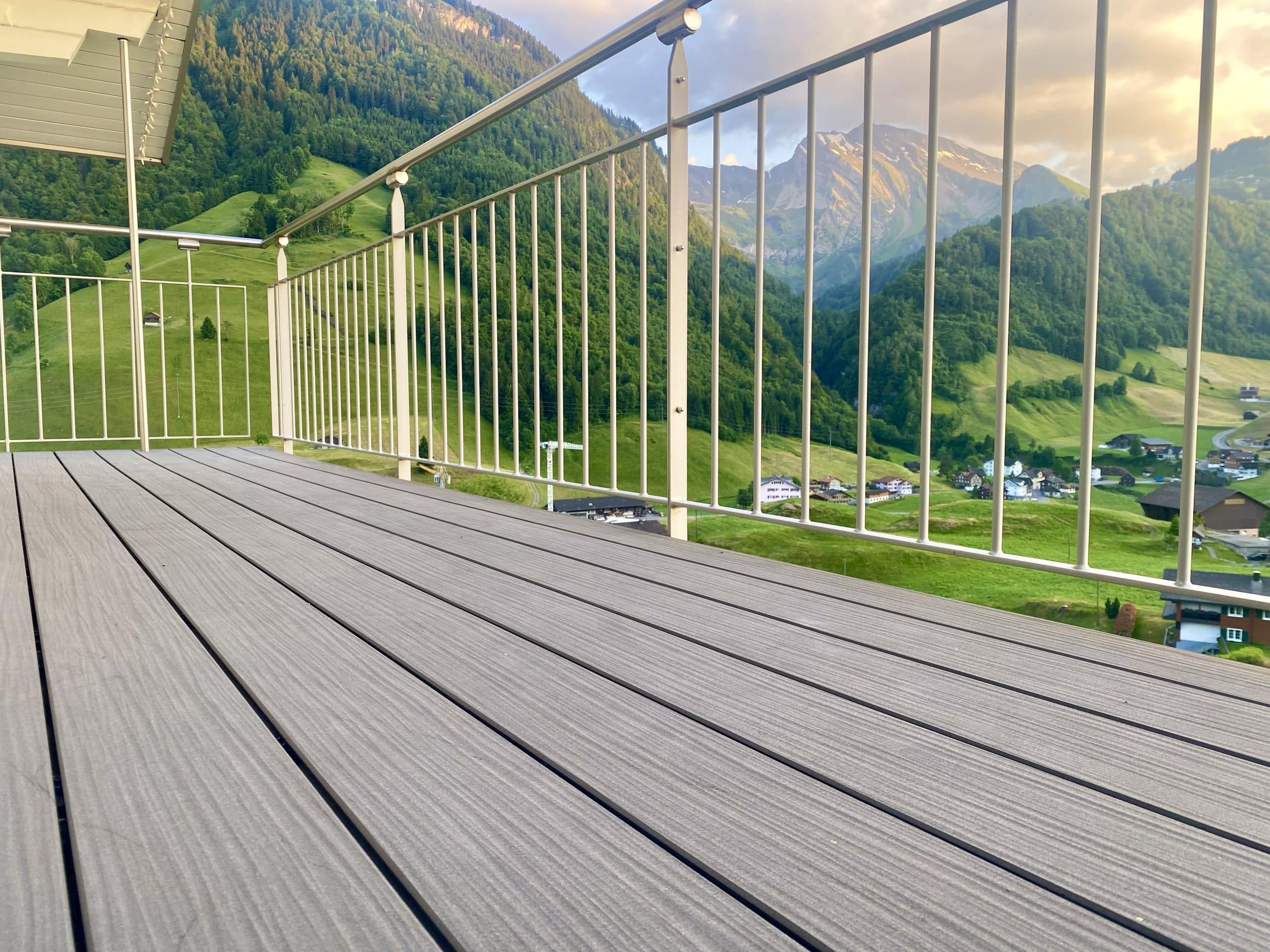 Ein Blick auf einen Balkon mit einem modernen Geländer, der eine malerische Aussicht auf grüne Hügel und Berge bietet. Der Boden ist aus grauem, strukturierter Holz- oder Verbundmaterial, das eine einladende Atmosphäre schafft.