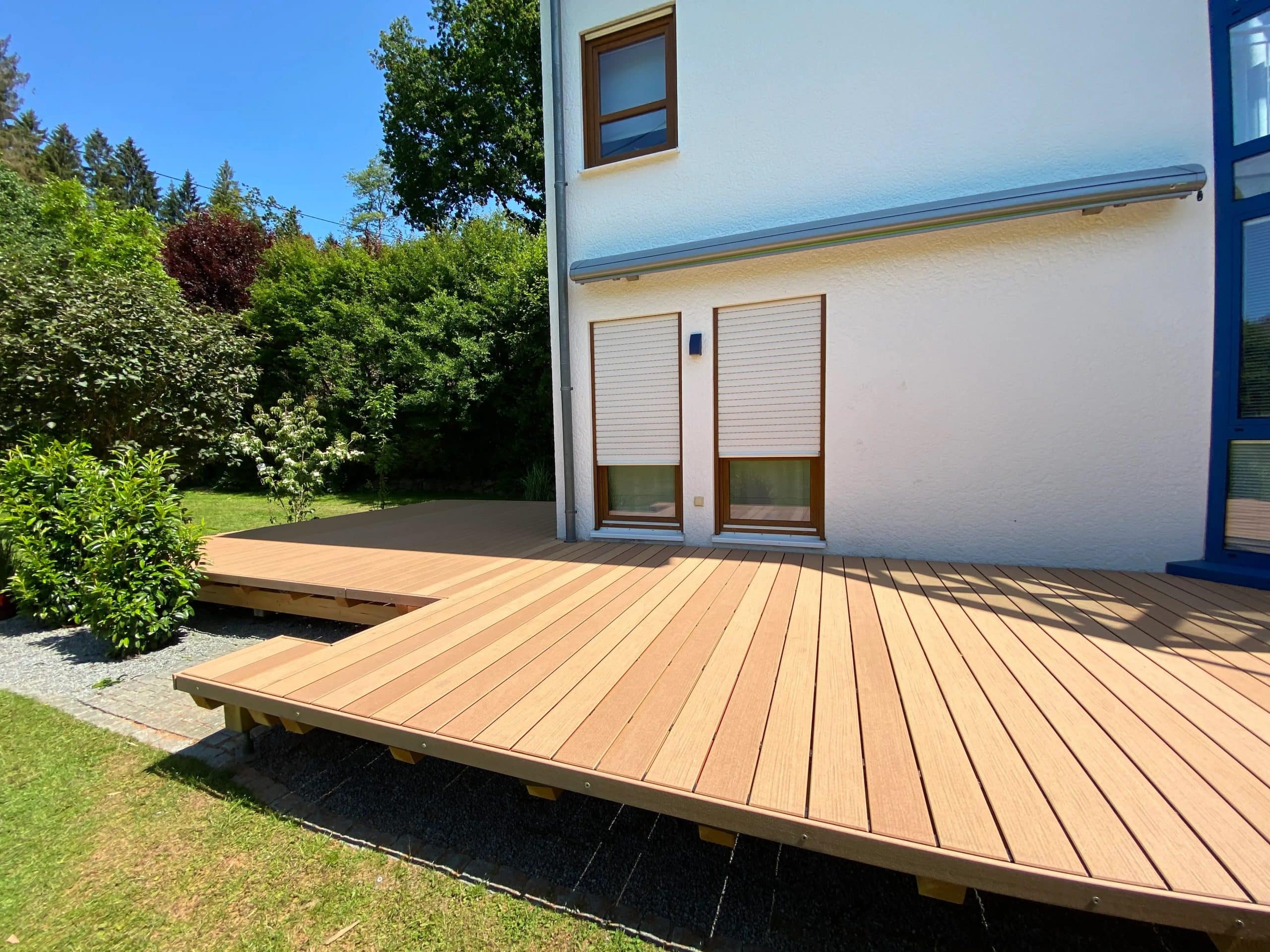 Terrasse aus Holz mit hellen Dielen, angrenzend an ein modernes Haus, umgeben von grünem Garten und Bäumen, bei sonnigem Wetter.