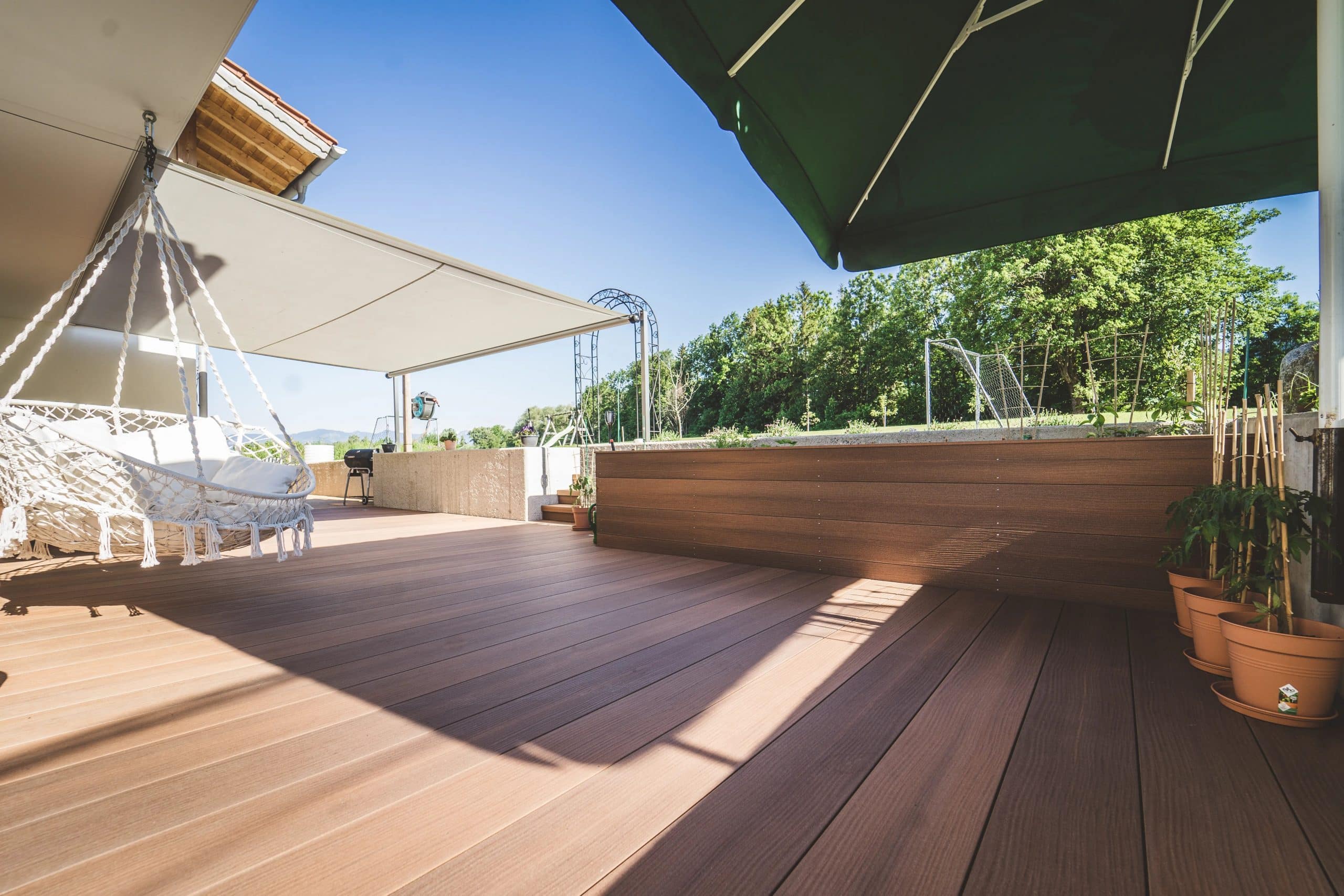 Ein sonniger Balkon mit Holzfußboden, einer weißen Hängematte und einem grünen Sonnenschirm. Im Hintergrund sind Bäume und ein Fußballtor sichtbar. Töpfe mit Pflanzen sind am Rand platziert.