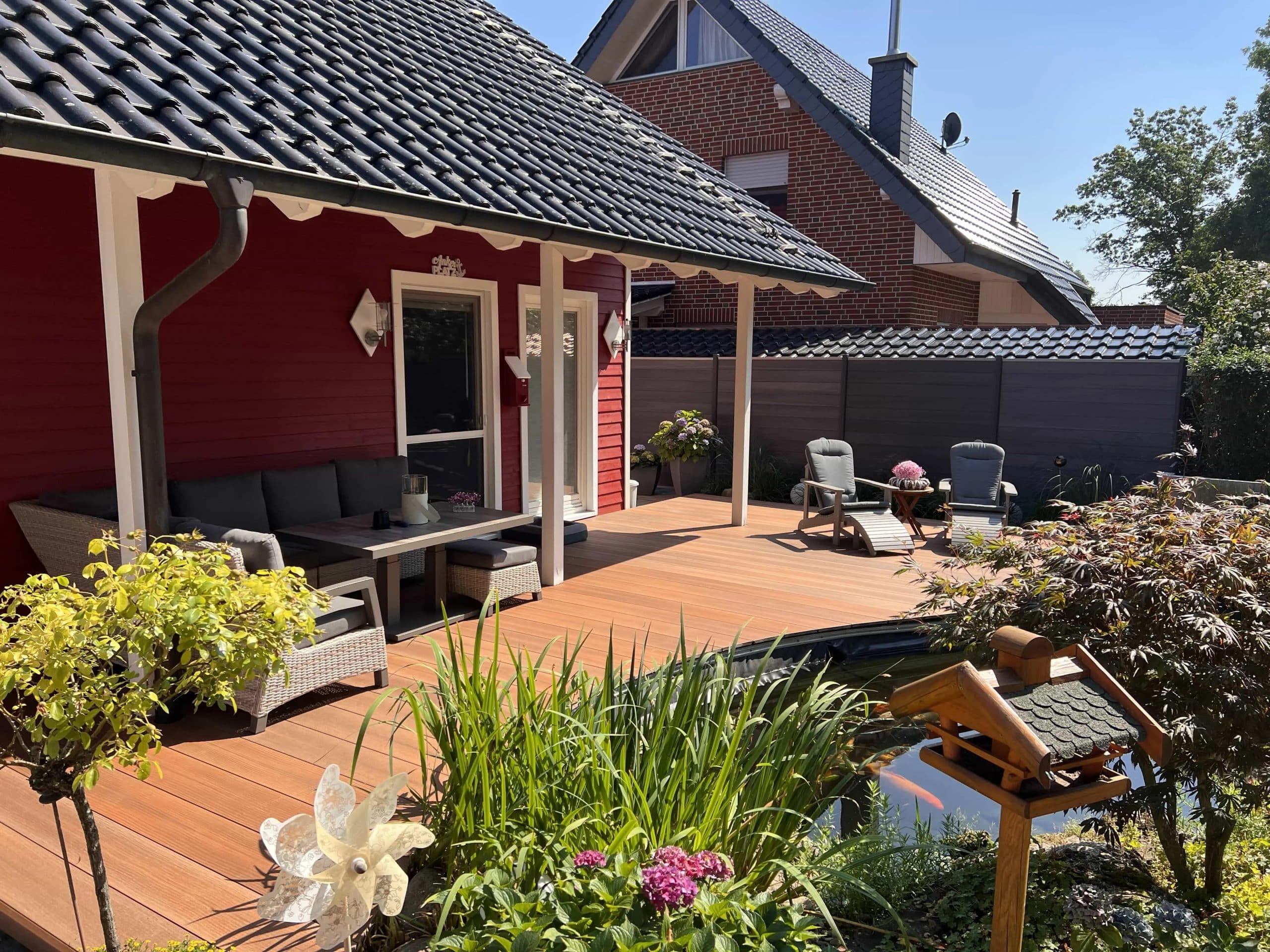 Ein gemütlicher Außenbereich mit einer Holzterrasse, einem grauen Sofa und einem Tisch. Im Hintergrund sind zwei Liegestühle und eine kleine Wasserstelle mit Pflanzen zu sehen. Die Wand des Hauses ist rot gestrichen, und der Himmel ist klar und blau.