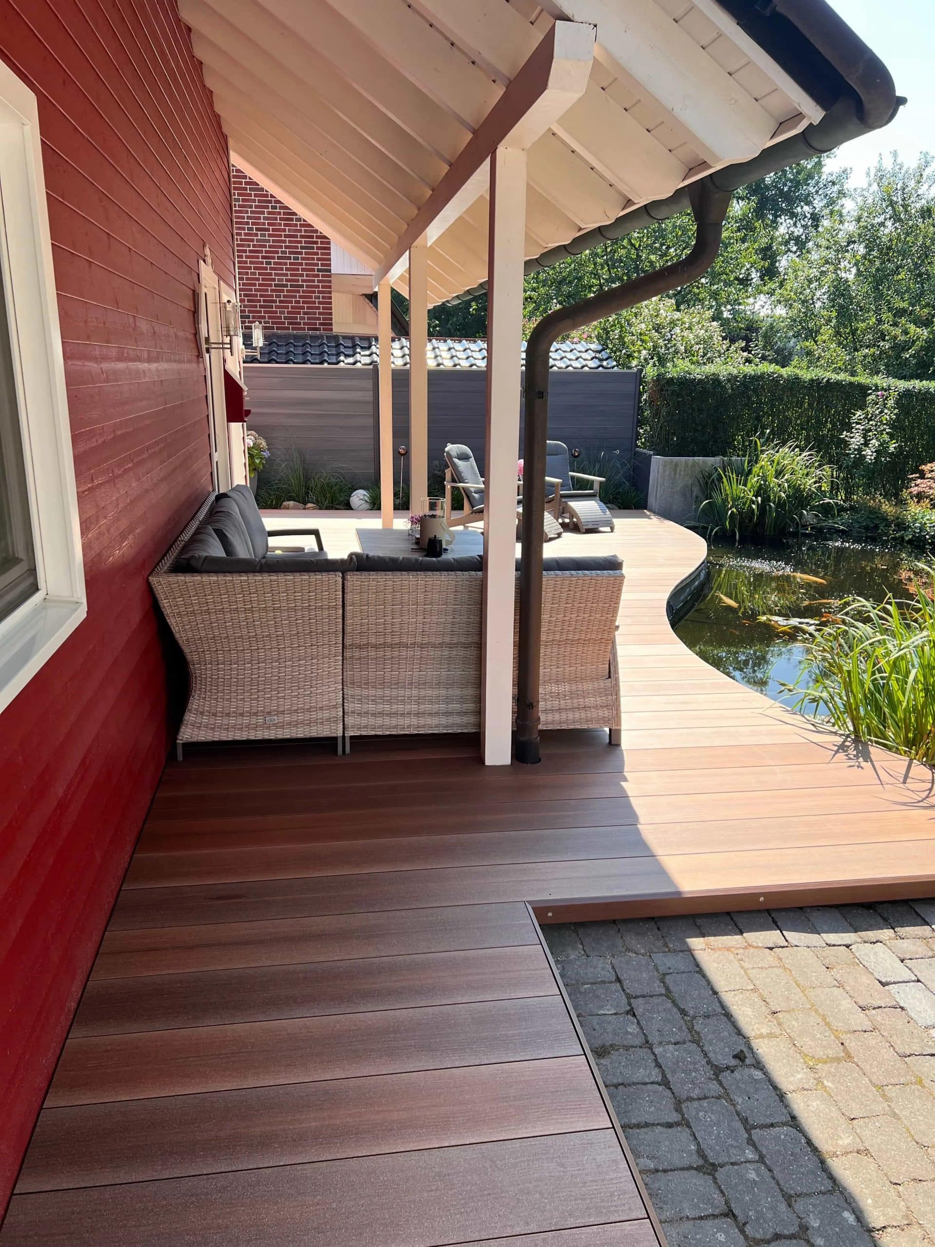 Eine Terrasse mit einem hellen Holzfußboden, einem gemütlichen Sitzbereich mit einem Rattan-Sofa und einem Blick auf einen kleinen Teich, umgeben von grünen Pflanzen und Sträuchern. Im Hintergrund ist ein rotes Holzhaus und ein gepflasterter Weg zu sehen.