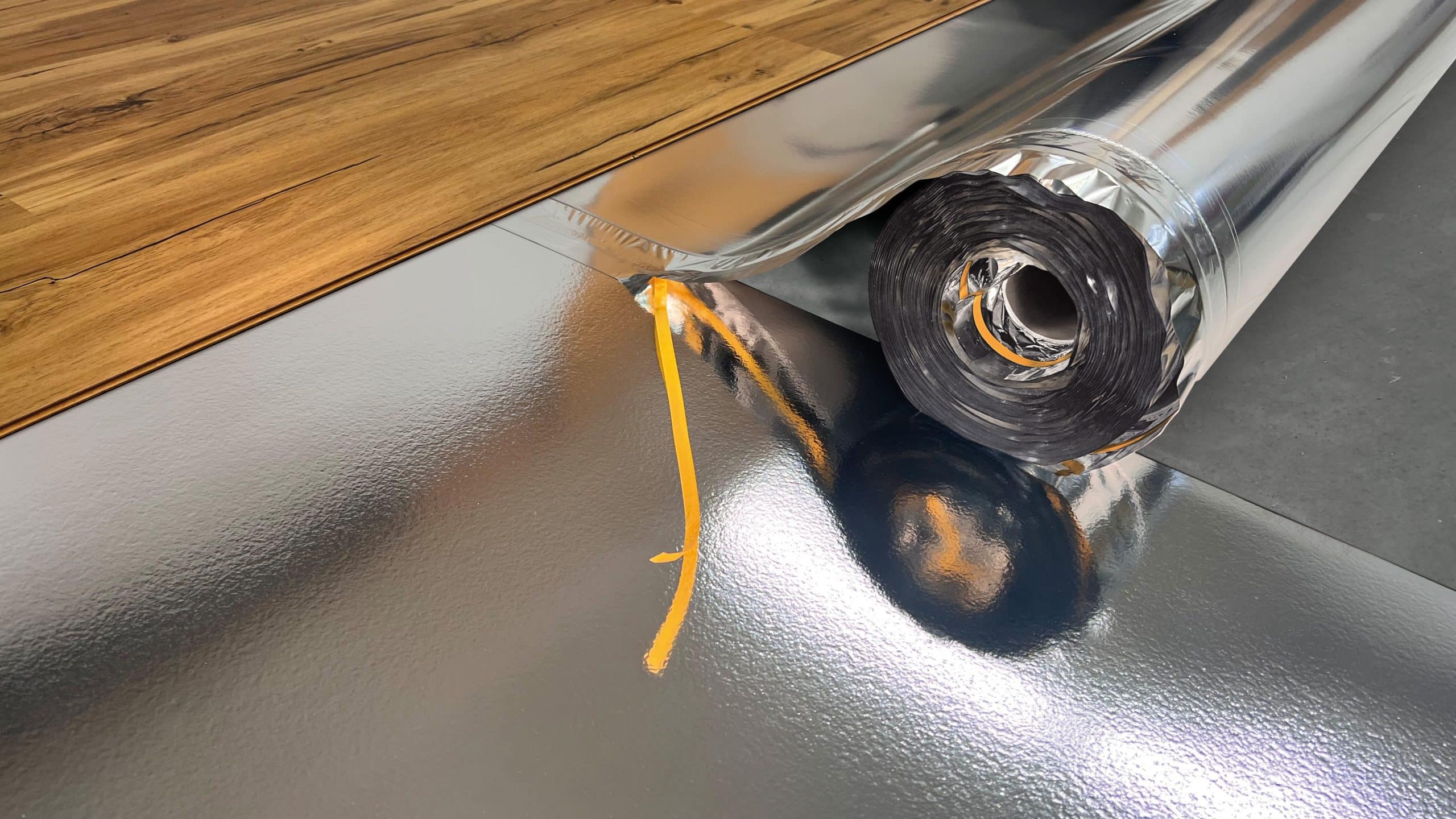 Eine silberne Folie liegt auf einem grauen Untergrund, während ein Rollenkern aus schwarzem Material sichtbar ist. Im Hintergrund ist ein Holzboden zu sehen, der einen warmen Farbton hat.