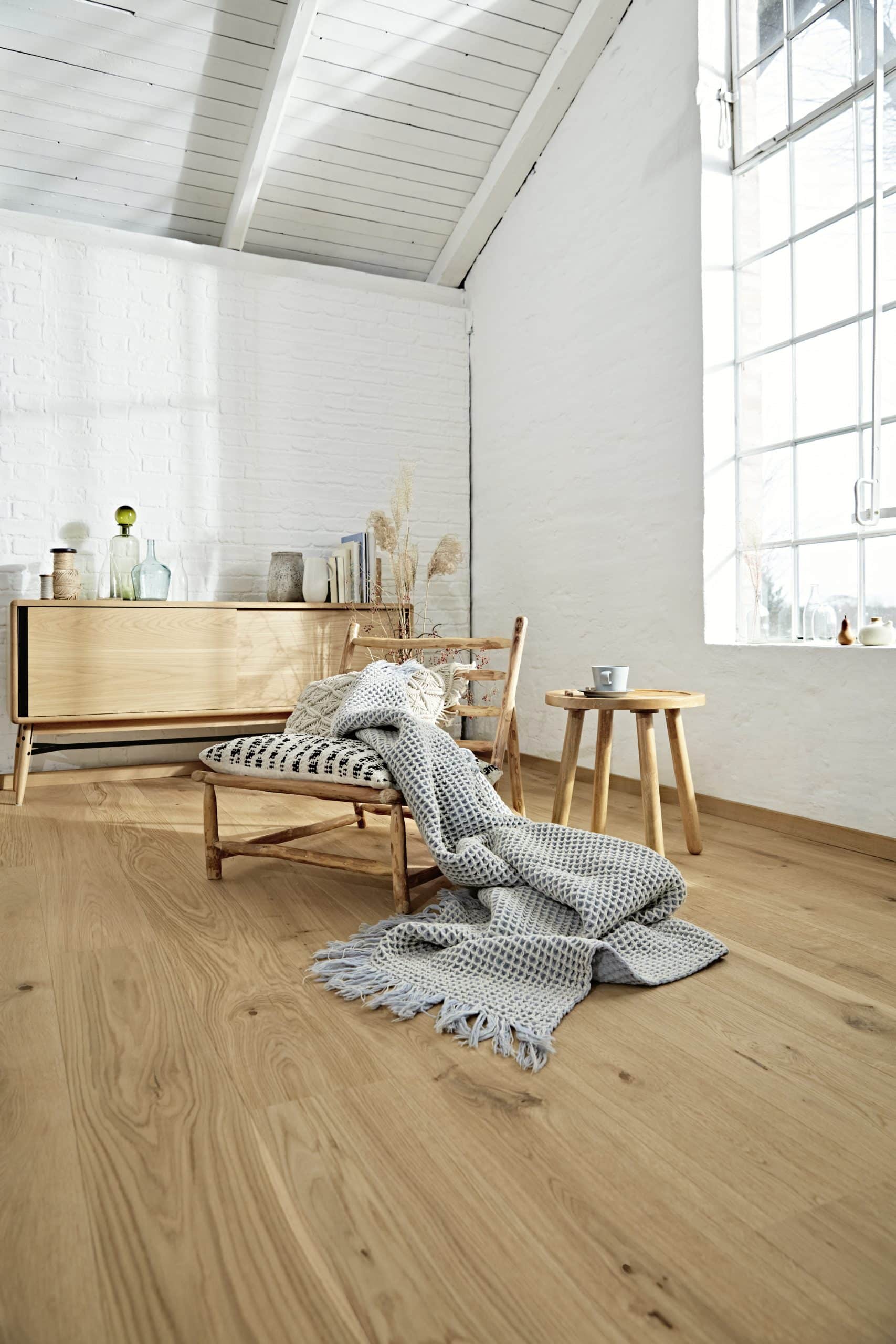 Ein gemütlicher Raum mit einem Holzstuhl, einer Decke und einem kleinen Tisch. An der Wand steht ein Sideboard mit dekorativen Flaschen und Büchern. Große Fenster lassen viel Tageslicht herein, und der Boden ist aus hellem Holz.