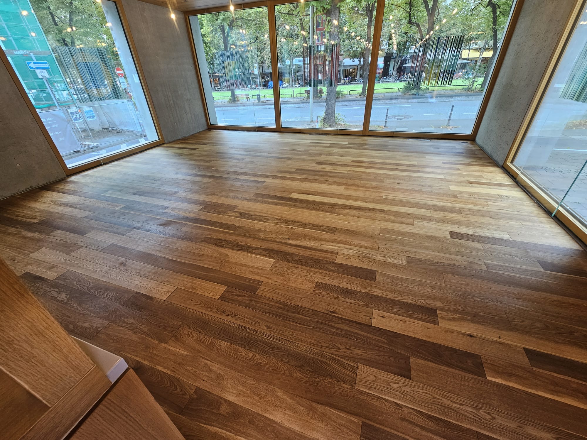 Ein heller Raum mit großen Fenstern, der mit einem modernen Holzboden ausgestattet ist. Die Wände sind aus Beton und der Raum wirkt einladend und lichtdurchflutet. Im Hintergrund sind Bäume und eine Straße sichtbar.