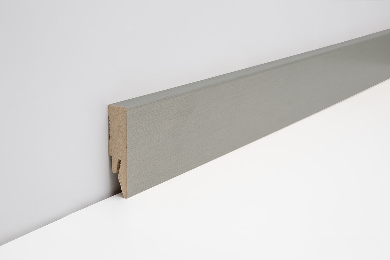 Eine graue Fußleiste aus MDF mit einer glatten, gebürsteten Oberfläche, die an einer Wand und einem weißen Boden montiert ist.