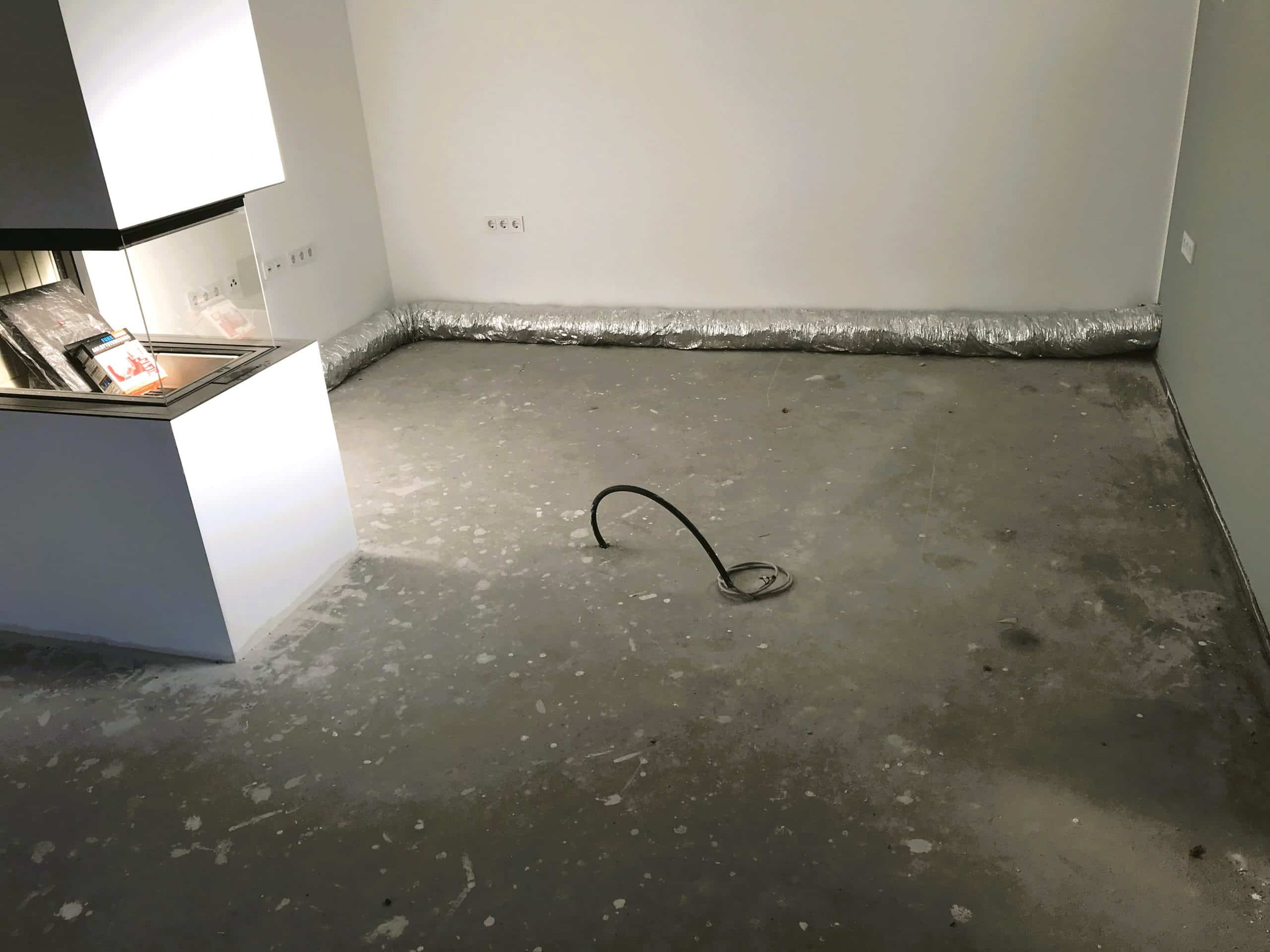 Ein leerer Raum mit einem grauen, unebenen Boden. An einer Wand verläuft eine silberne Isolierung, während in der Mitte des Raumes ein Kabel auf dem Boden liegt. Im Hintergrund ist ein Teil einer Küchenzeile sichtbar.
