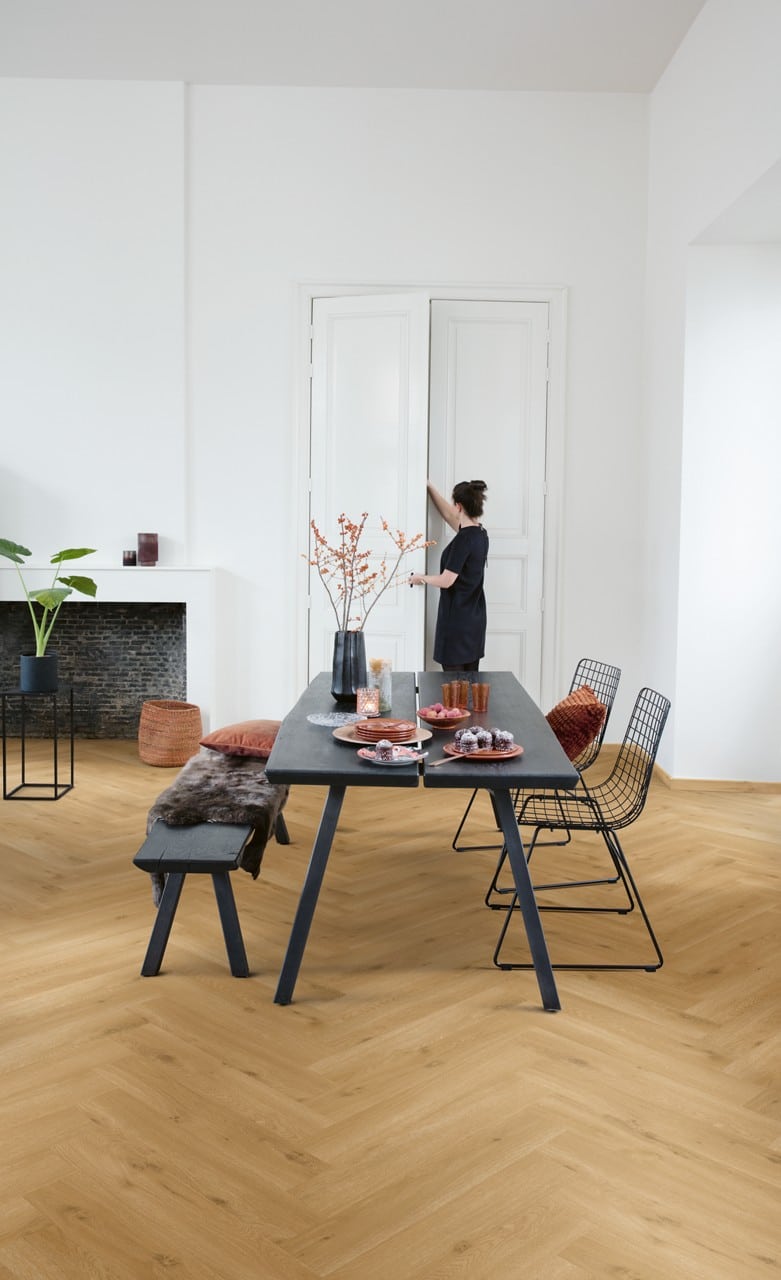 Ein moderner Essbereich mit einem schwarzen Tisch, Stühlen aus Draht und einer Bank. Auf dem Tisch sind Teller mit Snacks und eine Vase mit Zweigen. Im Hintergrund steht eine Person, die eine Tür öffnet, während eine grüne Pflanze und ein Kamin im Raum sichtbar sind. Der Boden besteht aus hellem Parkett.