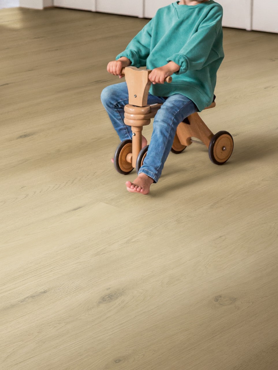 Ein kleines Kind sitzt auf einem Holzfahrzeug und fährt auf einem hellen, holzähnlichen Boden. Das Kind trägt einen grünen Pullover und Jeans, während es barfuß ist.