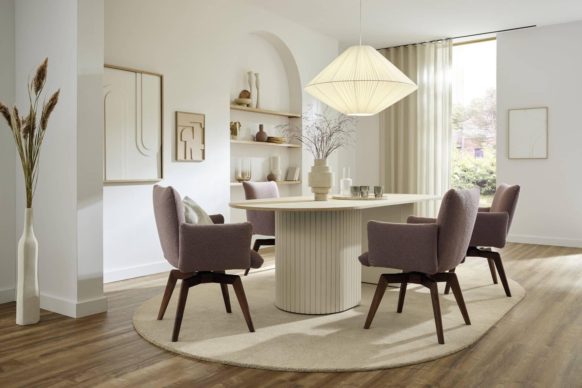 Ein modern gestalteter Essbereich mit einem runden Tisch, umgeben von gepolsterten Stühlen in sanften Farben. Eine große, geometrische Pendelleuchte hängt über dem Tisch. An einer Wand befinden sich dekorative Regale mit verschiedenen Vasen und Kunstwerken. Ein weicher Teppich und große Fenster mit Vorhängen tragen zur gemütlichen Atmosphäre bei.