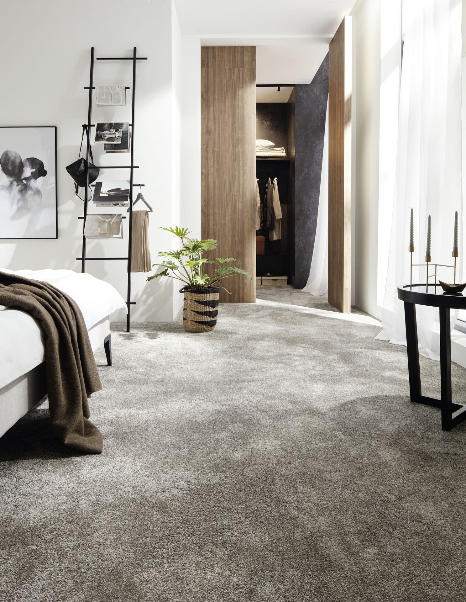 Ein modernes Schlafzimmer mit einem grauen Teppichboden, einem Bett mit einer braunen Decke, einer schwarzen Leiter zur Aufbewahrung von Kleidung und Accessoires, sowie einem großen Kleiderschrank, der durch eine Holzschiebetür zugänglich ist. Eine Pflanze und ein Tisch mit Kerzen ergänzen die Einrichtung.