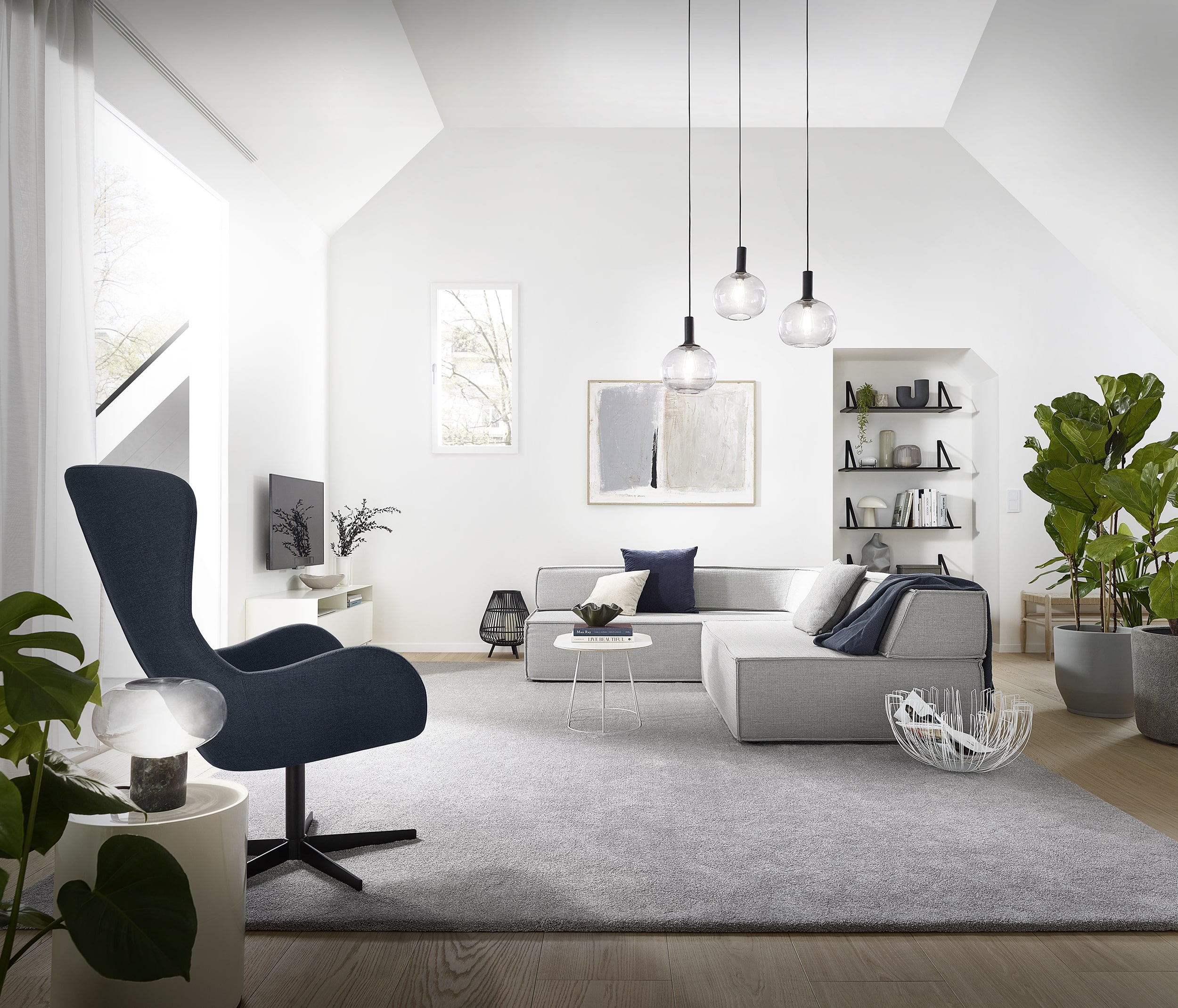 Heller, moderner Wohnraum mit schrägen Wänden, einem grauen Sofa, einem blauen Sessel und einem runden Tisch. An der Wand sind Bücherregale mit dekorativen Objekten und Pflanzen. Große Fenster lassen viel Tageslicht herein, während elegante Pendelleuchten den Raum beleuchten. Ein weicher Teppich sorgt für Gemütlichkeit.