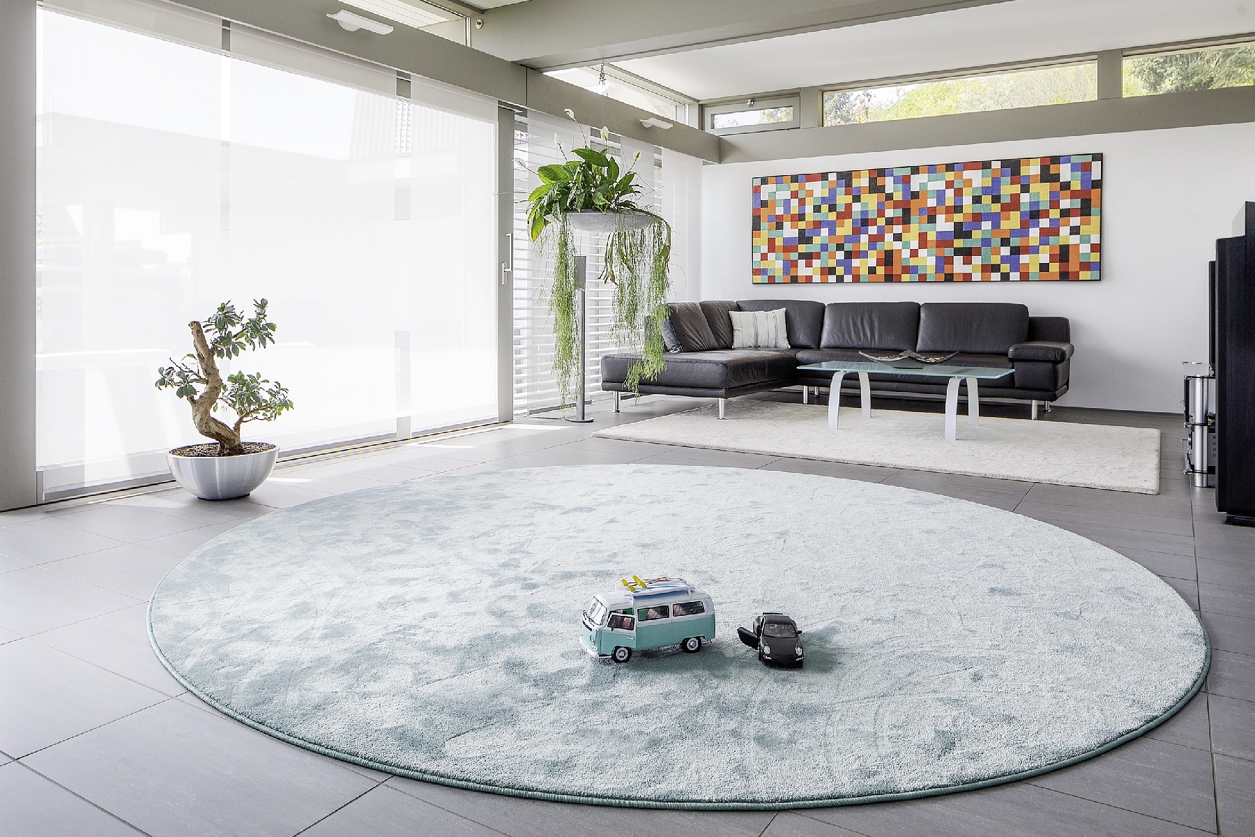 Ein moderner Wohnraum mit einem runden Teppich in sanften Farben, einem schwarzen Sofa, einem Glastisch und einer bunten Wandkunst. Im Vordergrund stehen zwei Spielzeugautos auf dem Teppich, während eine Zimmerpflanze in einem metallischen Topf zur Einrichtung beiträgt. Große Fenster sorgen für viel Tageslicht.