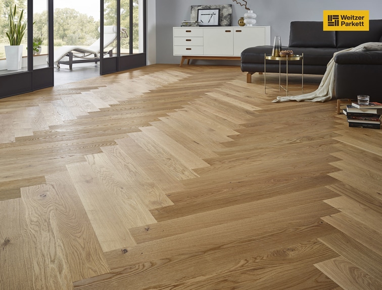 Ein eleganter Raum mit einem stilvollen Fischgrätenparkettboden aus hellem Holz. Im Hintergrund sind große Fenster, die viel Tageslicht hereinlassen, und ein modernes Wohnzimmer mit einem schwarzen Sofa und einem Couchtisch zu sehen. Ein weißer Schrank mit dekorativen Elementen ergänzt die Einrichtung.