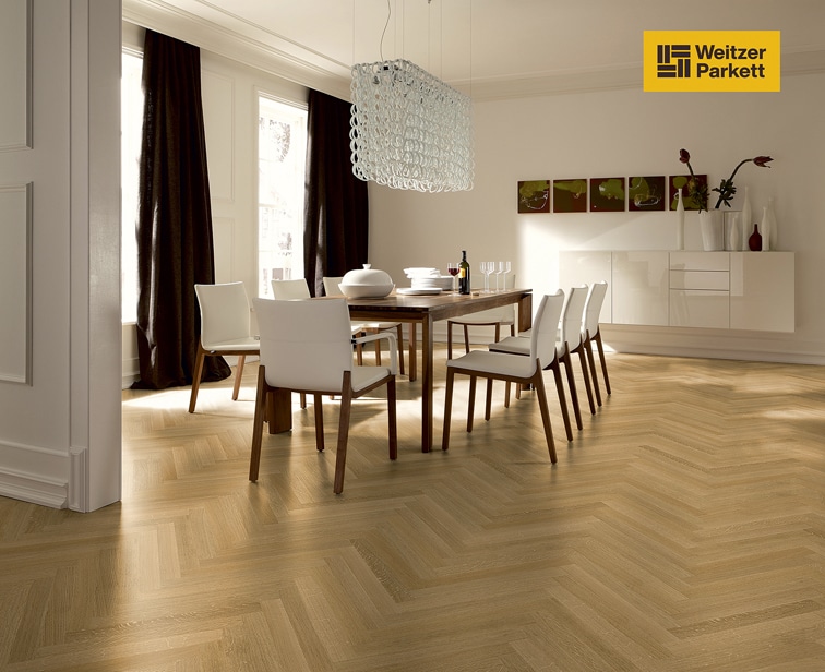 Ein modern eingerichteter Essbereich mit einem Holztisch, umgeben von weißen Stühlen. Über dem Tisch hängt eine stilvolle, gläserne Pendelleuchte. Der Boden ist mit hellen Parkettfliesen im Fischgrätmuster ausgelegt. An der Wand sind dunkle Vorhänge und ein elegenes Sideboard sichtbar.
