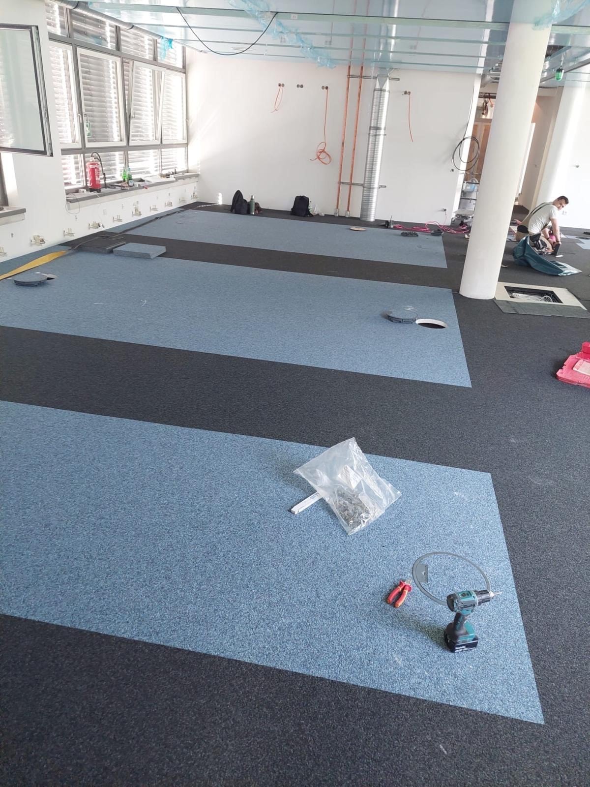 Ein leerer Raum mit einem teilweise verlegten Teppichboden in Blau- und Grautönen. Werkzeuge und Materialien liegen auf dem Boden, darunter eine Tüte mit Schrauben und ein Akku-Schrauber. Im Hintergrund sind Fenster und einige Baugeräte sichtbar.