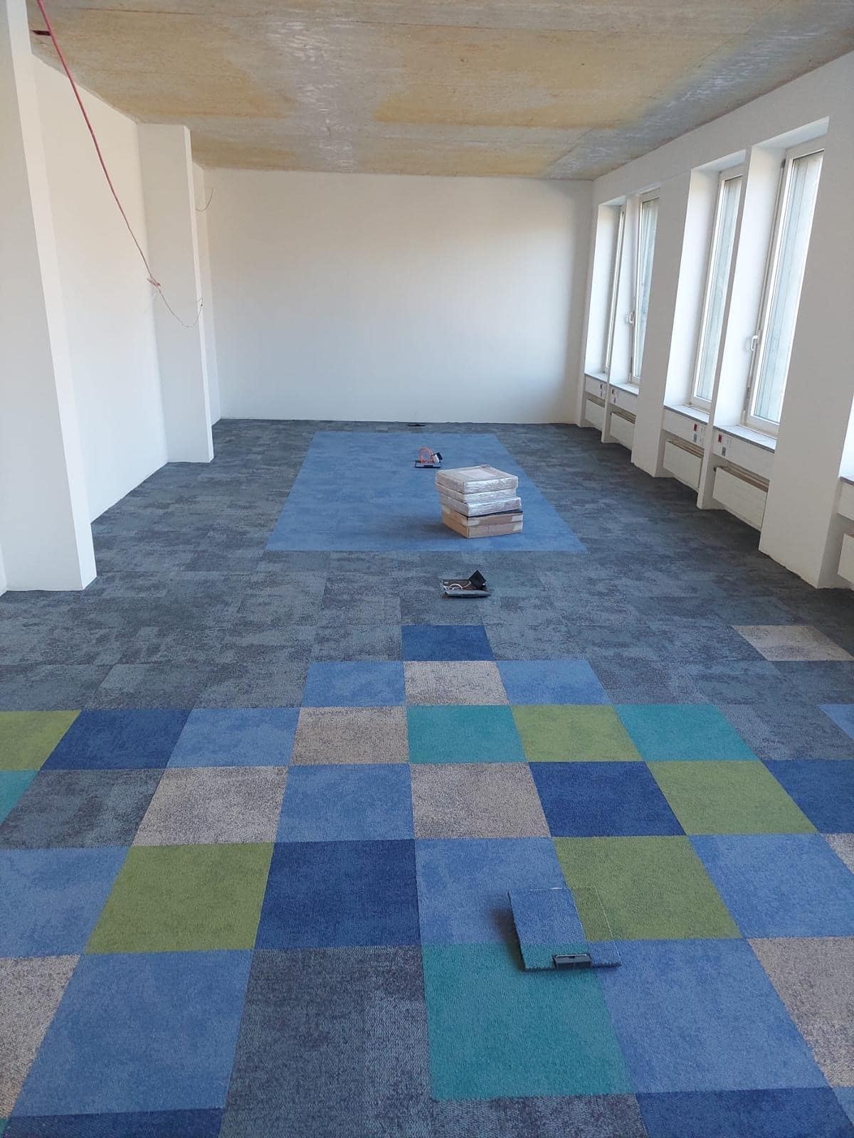 Ein leerer Raum mit hellen Wänden und großen Fenstern. Der Boden ist teilweise mit bunten Teppichfliesen in Blau-, Grün- und Grautönen ausgelegt. In der Mitte des Raumes liegen einige Stapel von Teppichfliesen, und ein paar lose Fliesen sind auf dem Boden verstreut. An der Decke sind Kabel sichtbar.