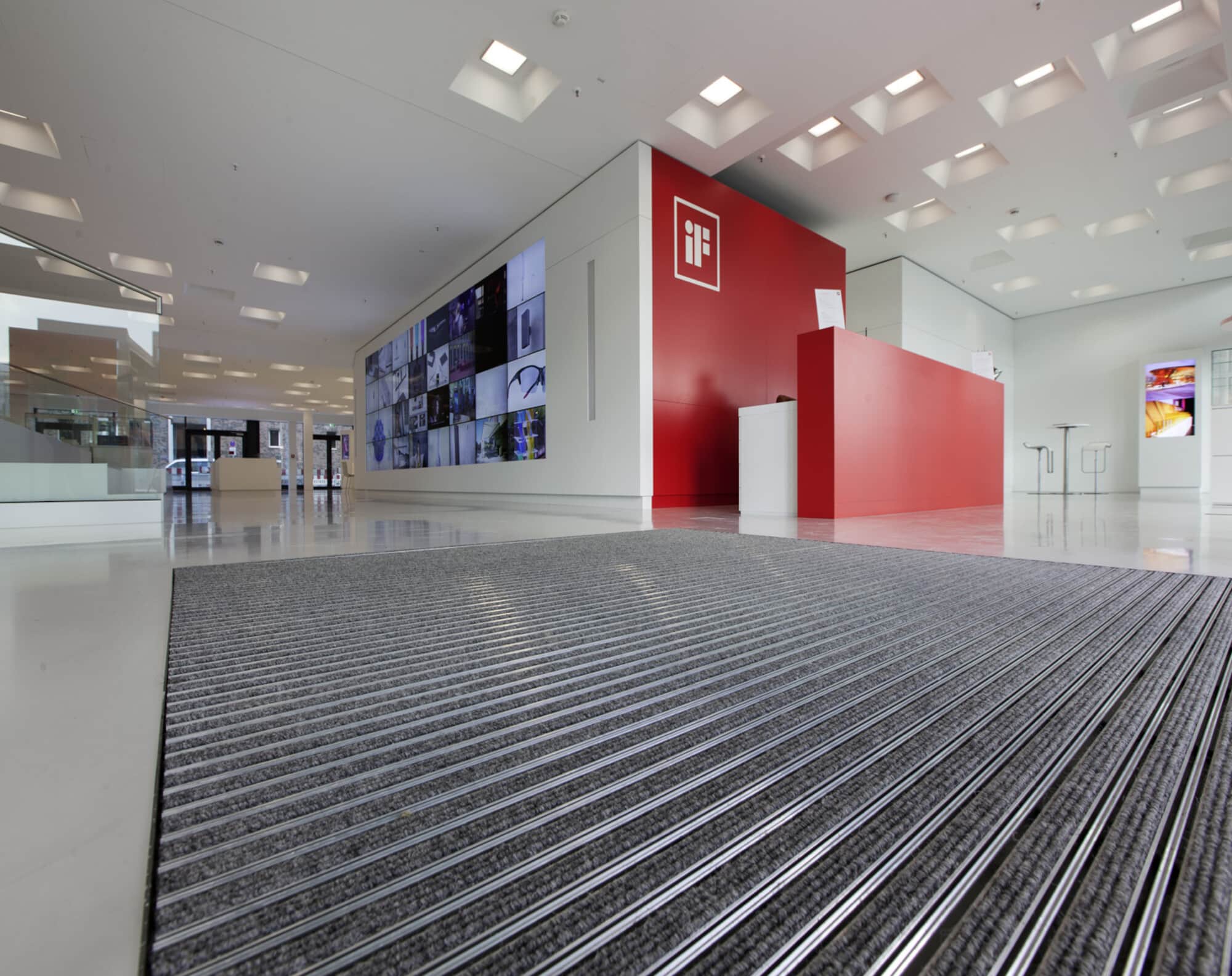 Moderne Empfangshalle mit einem roten Empfangstresen, einem großen Bildschirm mit verschiedenen Bildern an der Wand und einem sauberen, eleganten Bodenbelag.