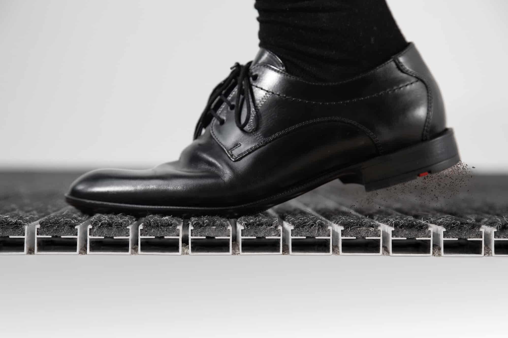 Ein schwarzer, eleganter Schuh tritt auf eine spezielle Bodenmatte, aus der Staubpartikel aufwirbeln.