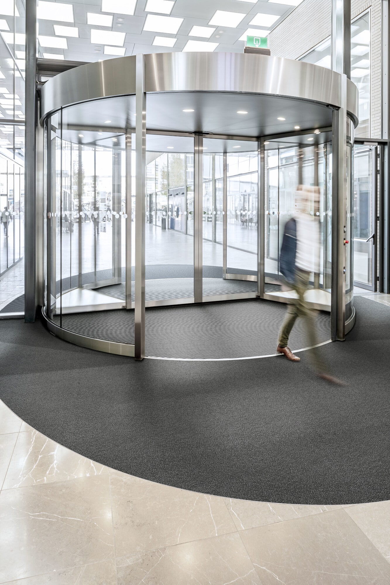 Ein moderner, runder Drehtür-Eingang in einem hellen, eleganten Gebäude. Die Tür besteht aus Glas und Edelstahl, während der Boden aus einem dunklen, strukturierten Material und hellen Fliesen besteht. Eine Person geht schnell durch die Drehtür.