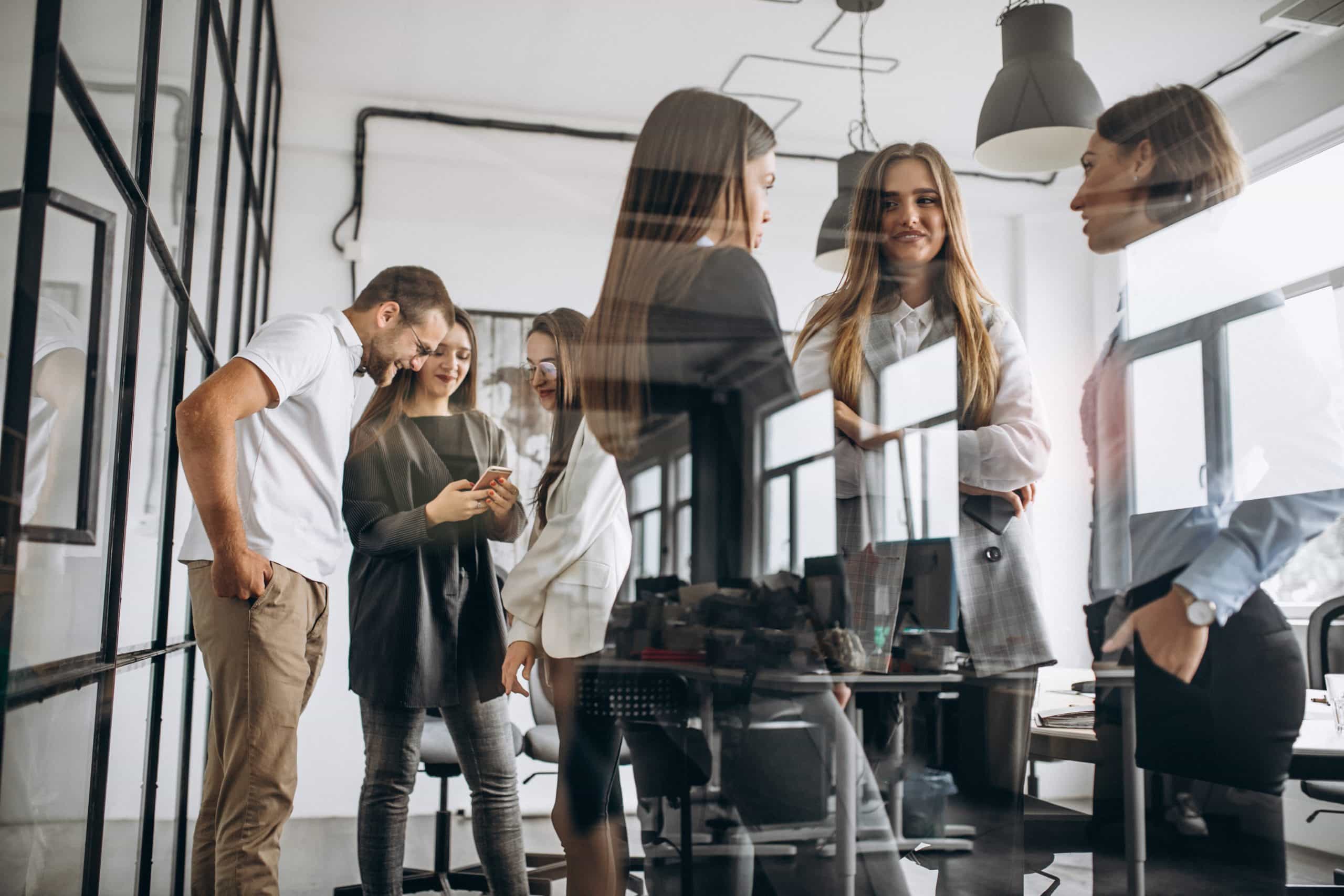 Eine Gruppe von fünf jungen Berufstätigen in einem modernen Büro, die miteinander sprechen und lachen. Zwei Frauen stehen im Vordergrund und betrachten gemeinsam ein Handy, während die anderen drei in einem Gespräch vertieft sind. Die Glasscheiben des Büros reflektieren das Geschehen und schaffen eine dynamische Atmosphäre.